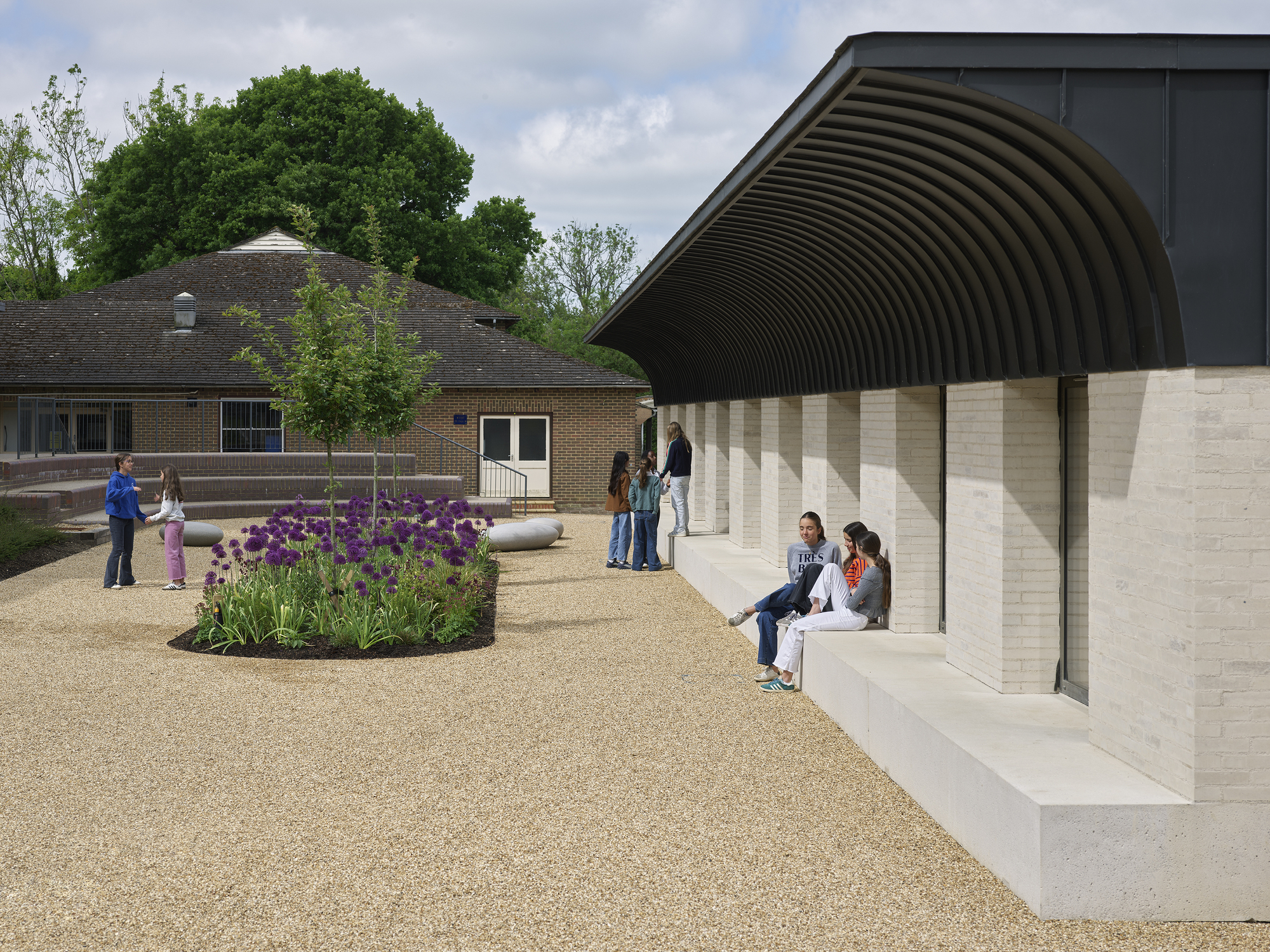 Courtyard with gravel surface and planted beds beside the ribbed metal canopy where visitors gather
