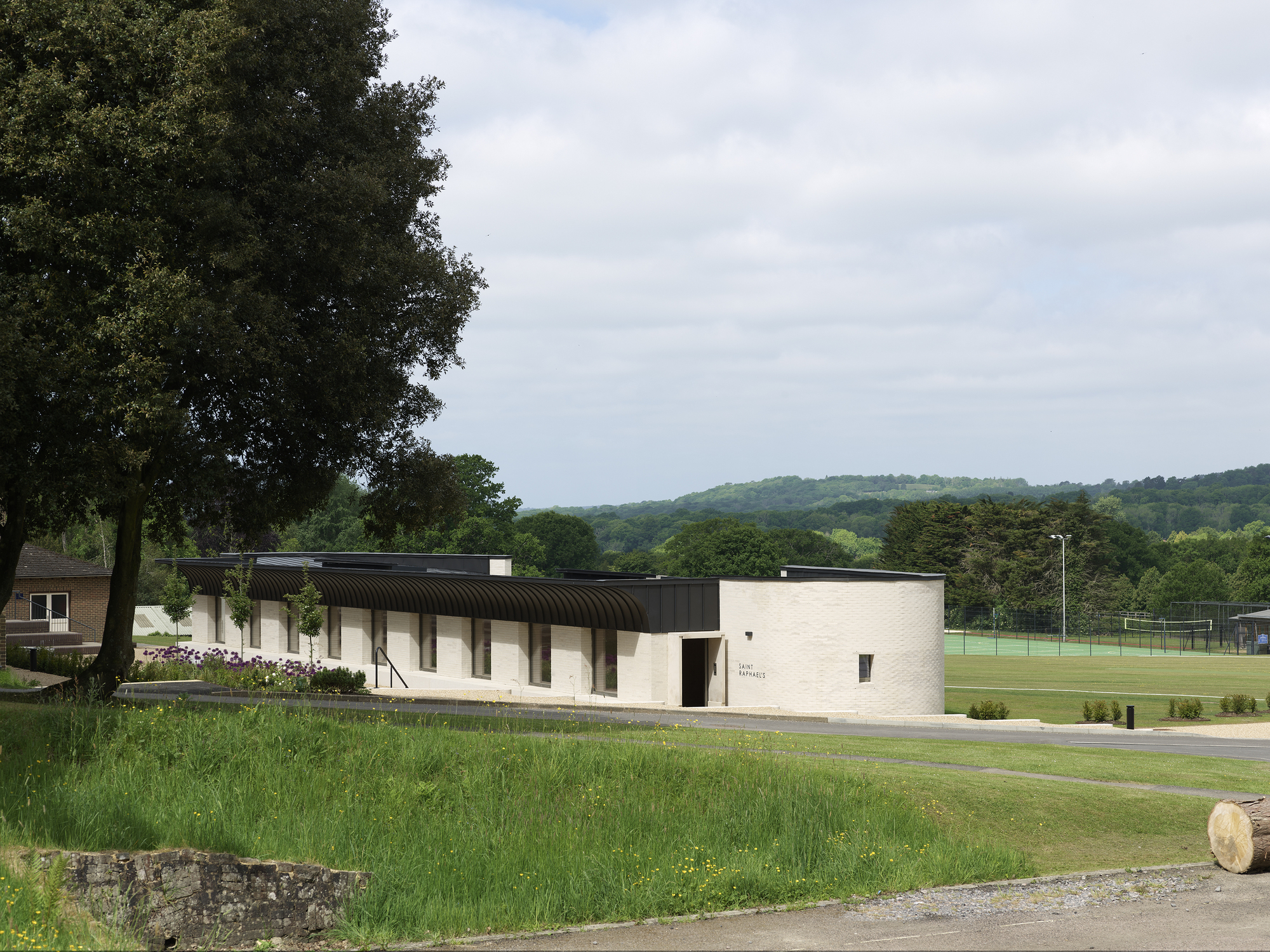 Low-rise pavilion with corrugated barrel-vault roof and pale brick walls set beside a sports field