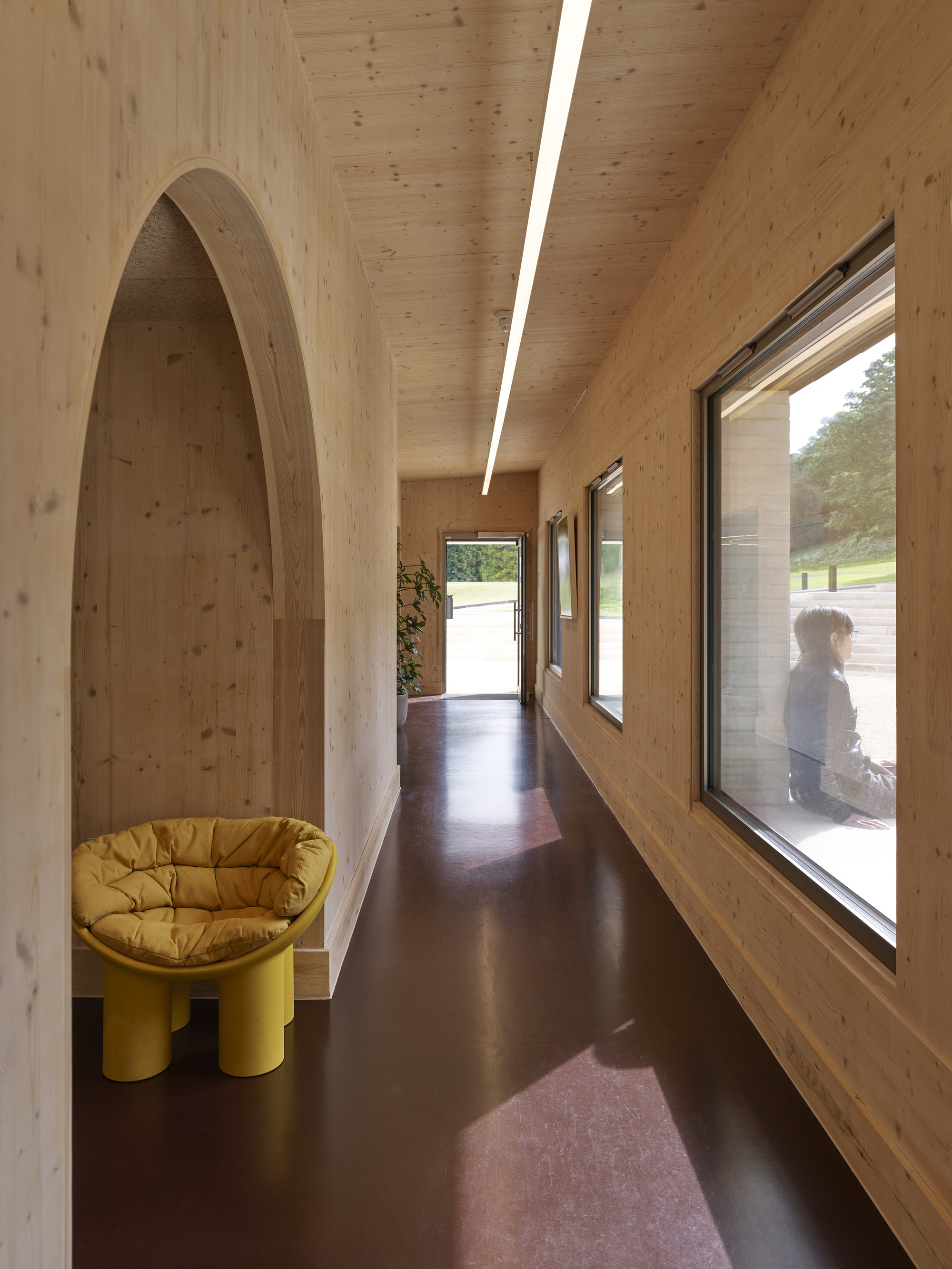 Arched timber corridor with skylight and yellow chair beside a window overlooking a person outside