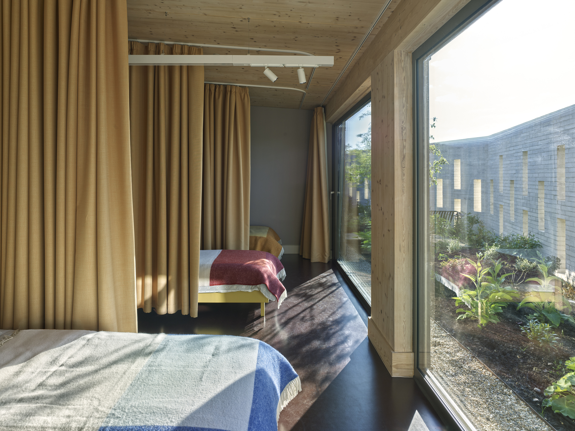 Bedroom with full-height glazing overlooking a planted courtyard framed by timber beams and ochre curtains