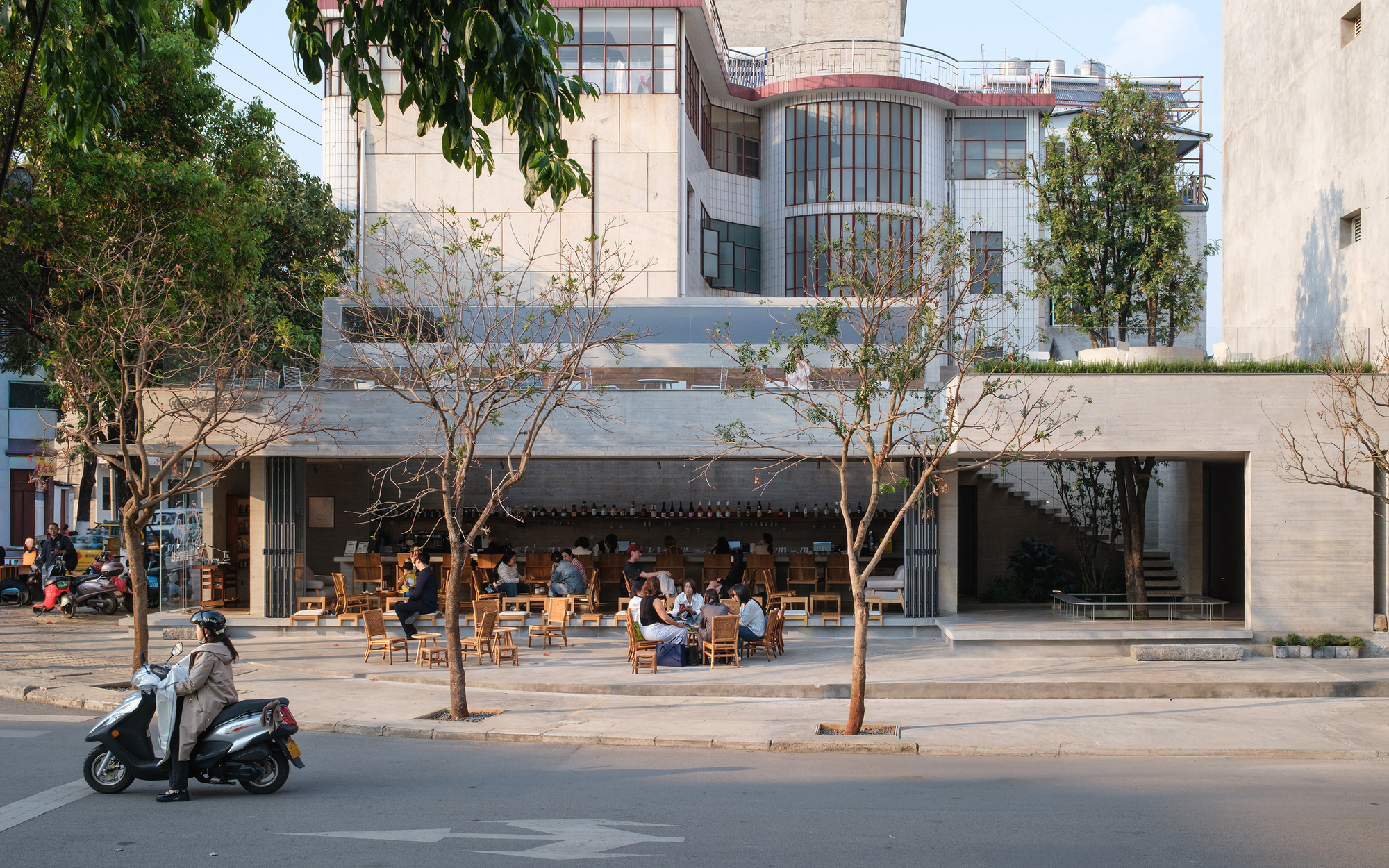 Street view of a cafe with open facade and outdoor seating under street trees with a motorbike passing