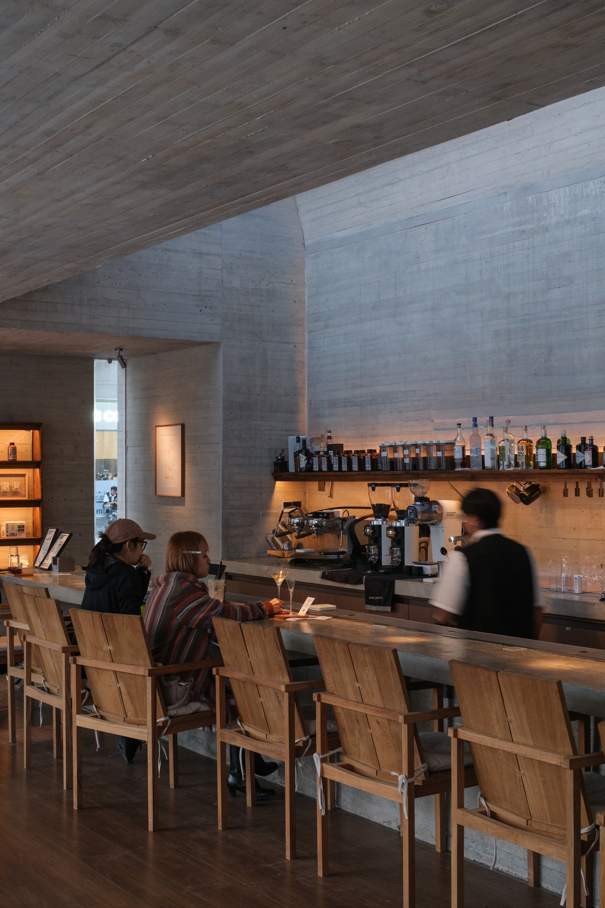 Cafe counter with patrons seated on wooden chairs beneath angled board-formed concrete ceiling at dusk