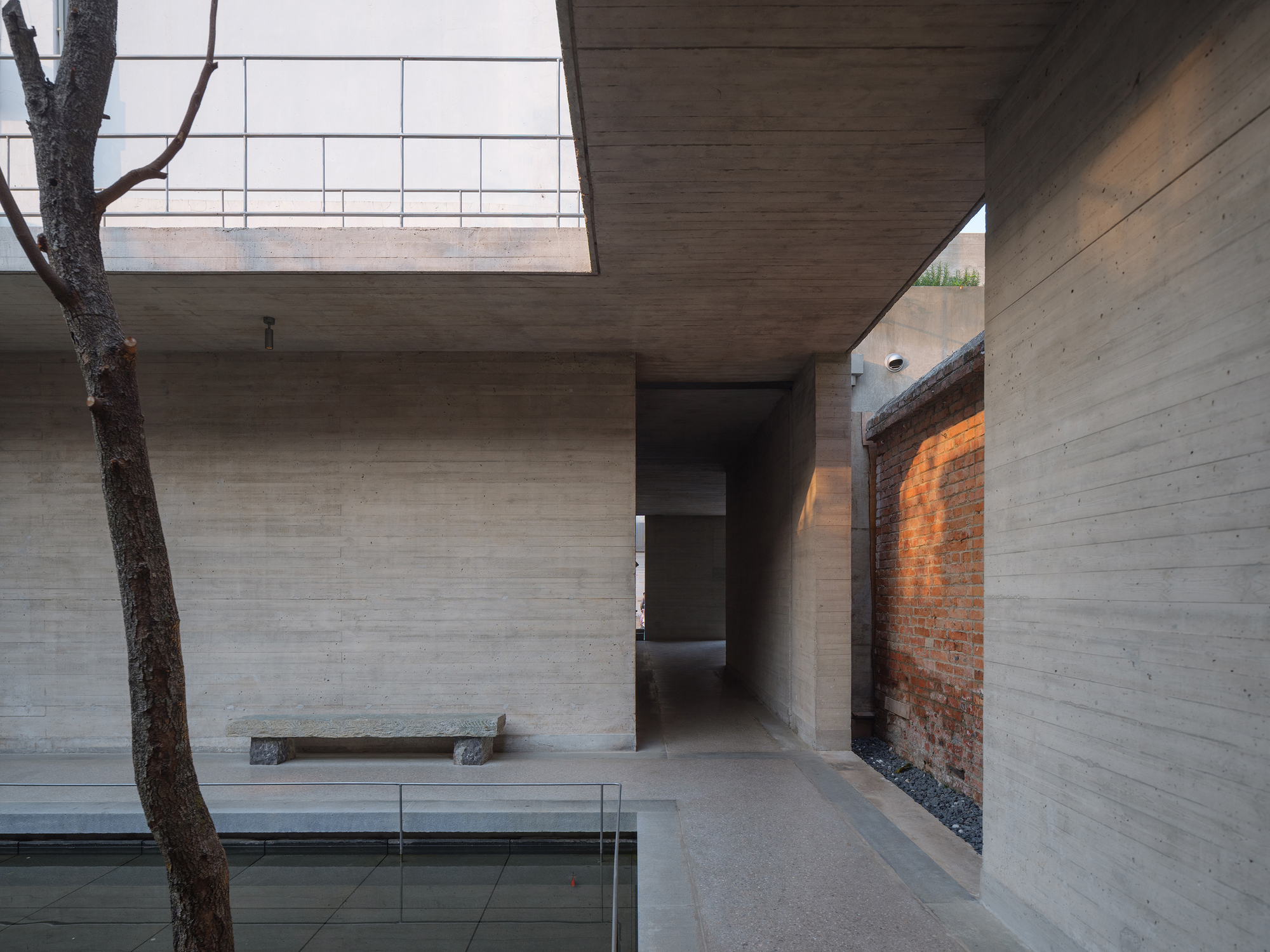 Courtyard view showing board-formed concrete walls, a tree trunk, and exposed brick remnant wall in afternoon light