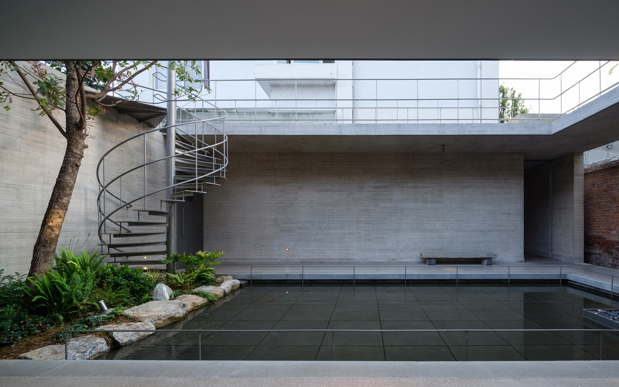 Concrete courtyard with reflecting pool, spiral staircase, and planted bed with rocks beneath an overcast sky