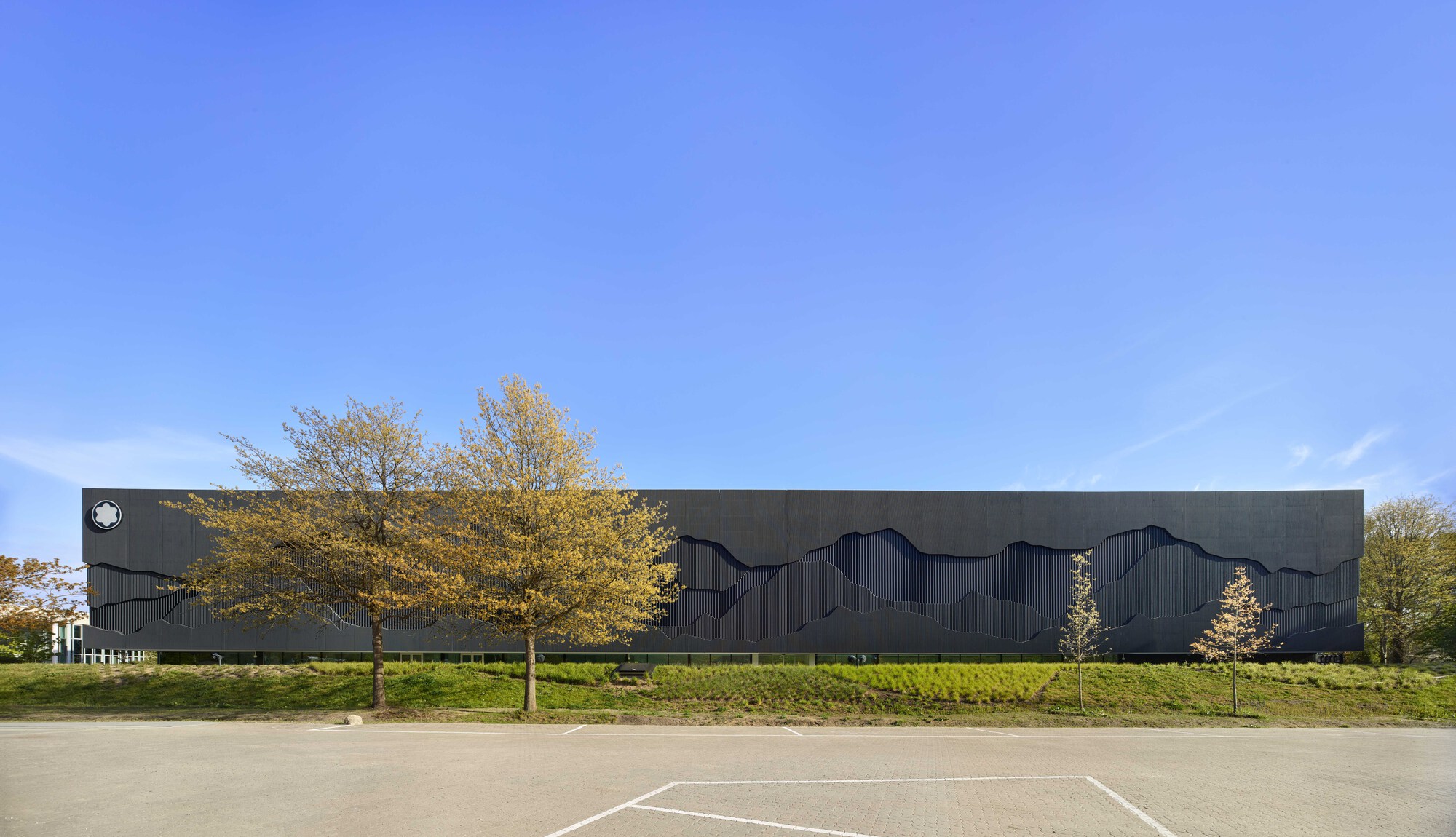 Dark metal facade with undulating relief pattern set behind a hedge and autumn trees