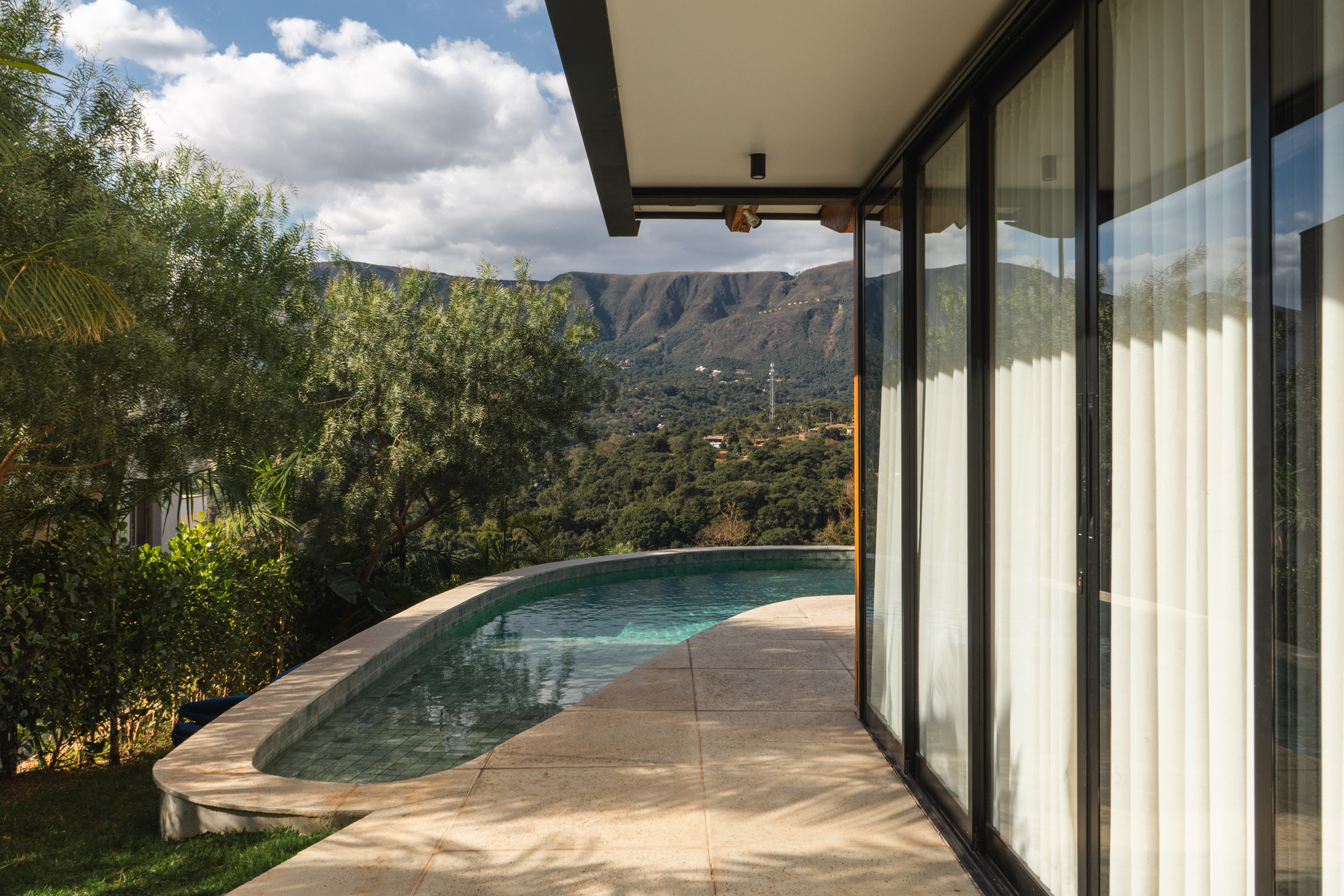 Curved pool terrace with stone pavers beside full-height sliding glass walls and mountain backdrop