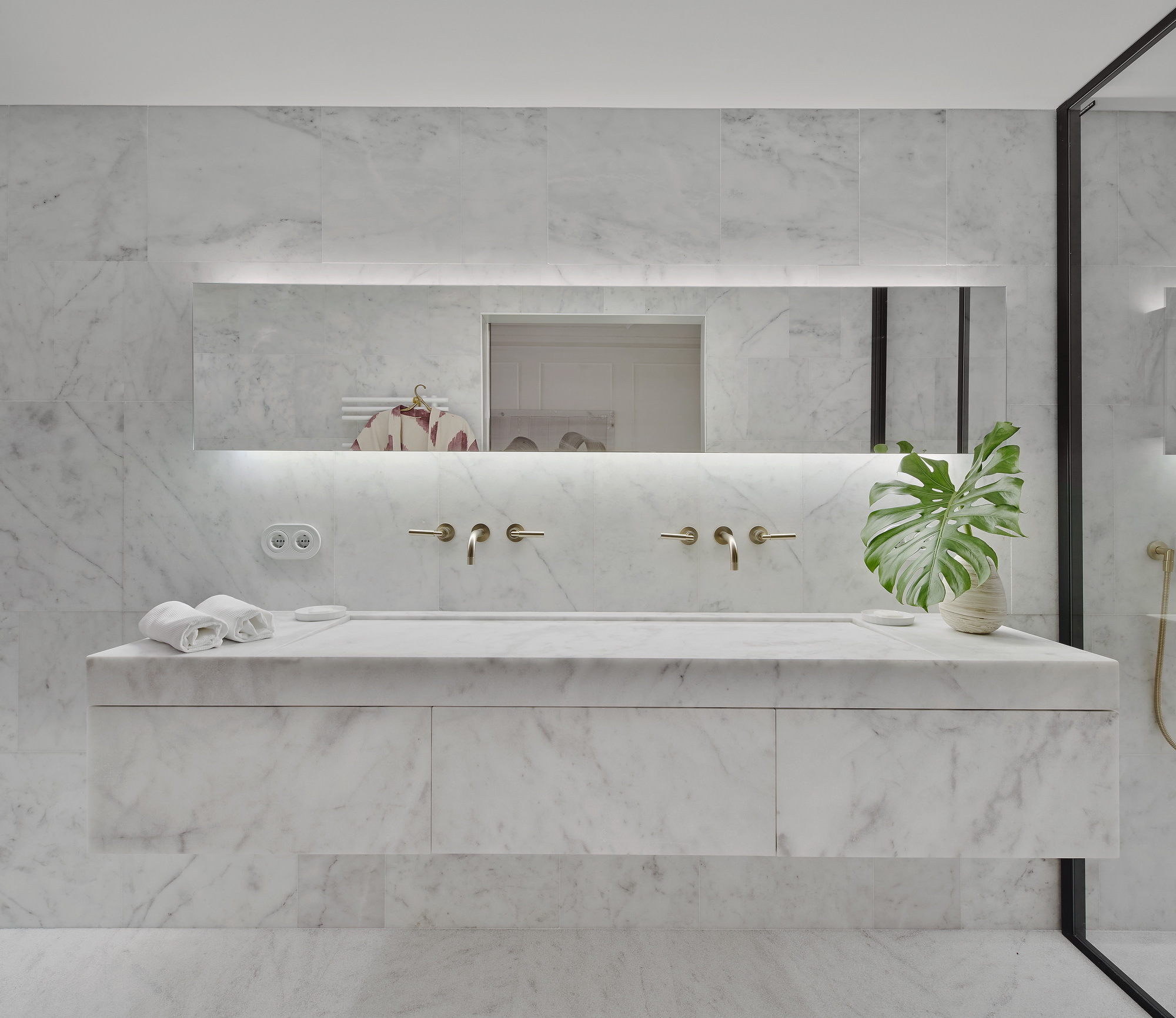 White marble vanity with integrated trough sink and brass wall-mounted faucets beneath a backlit mirror