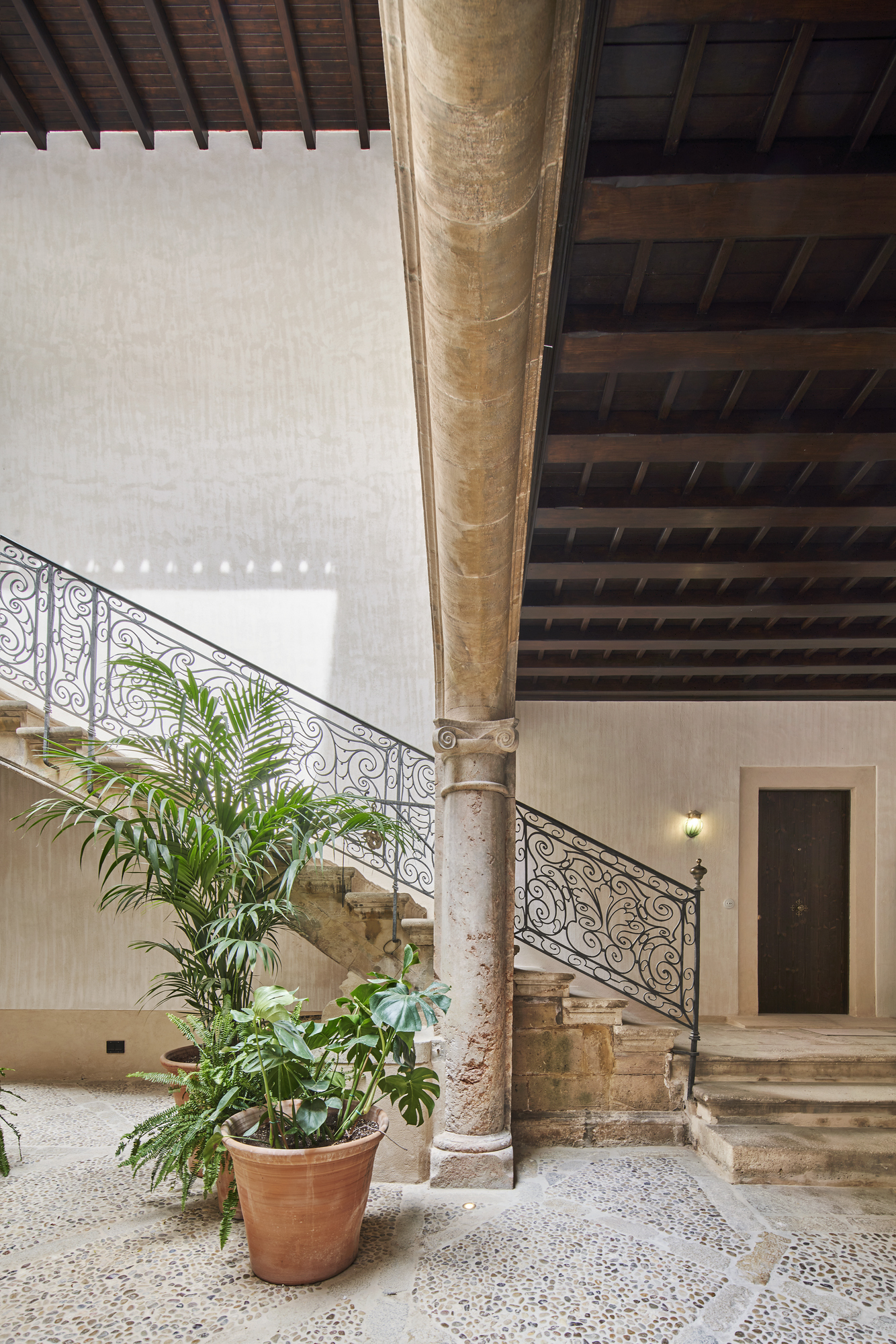 Stone staircase with wrought iron railing beside a weathered column and potted palm in a terrazzo courtyard