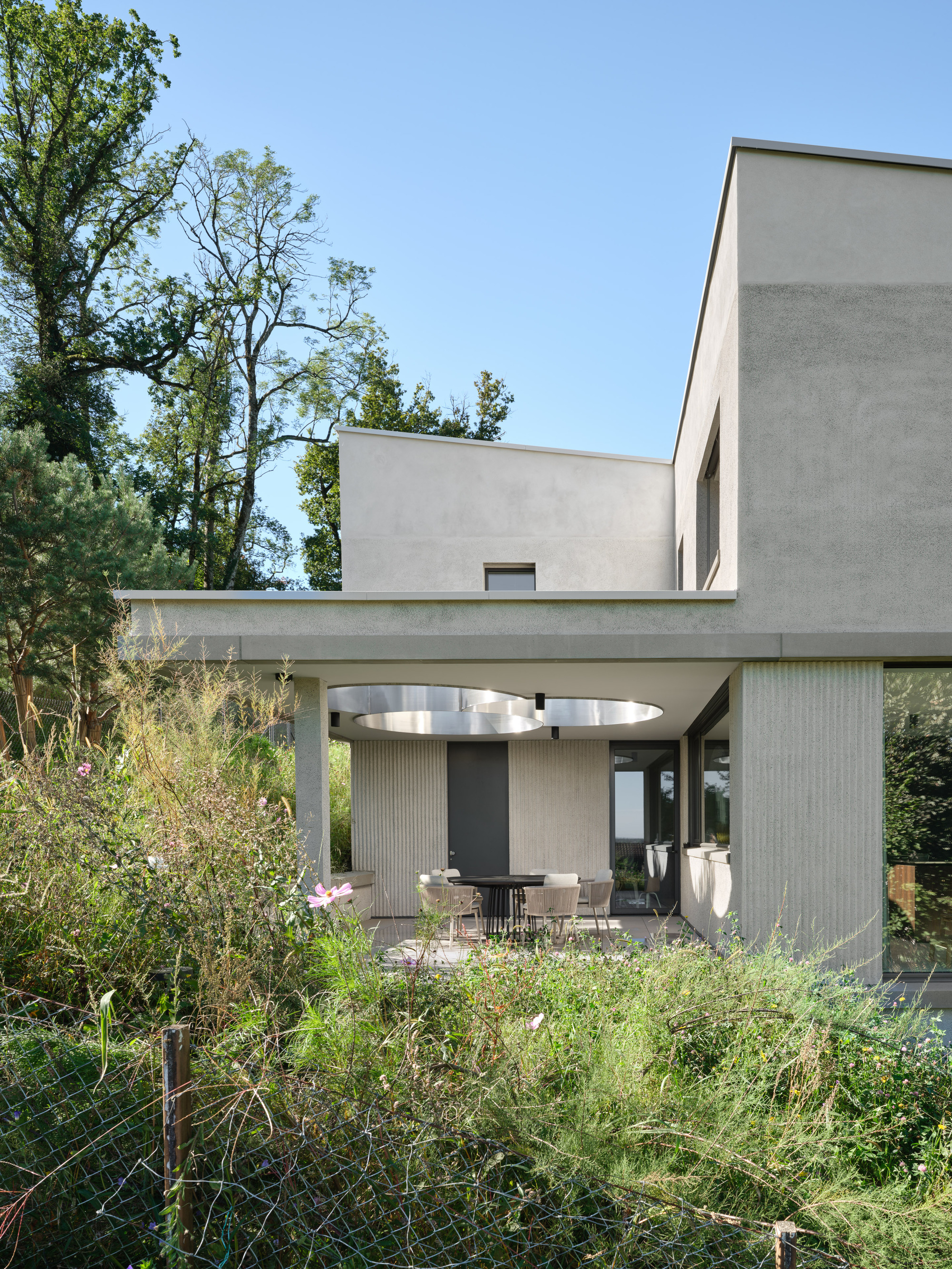 Covered outdoor terrace with concrete frame and circular skylight opening above a dining area in daylight