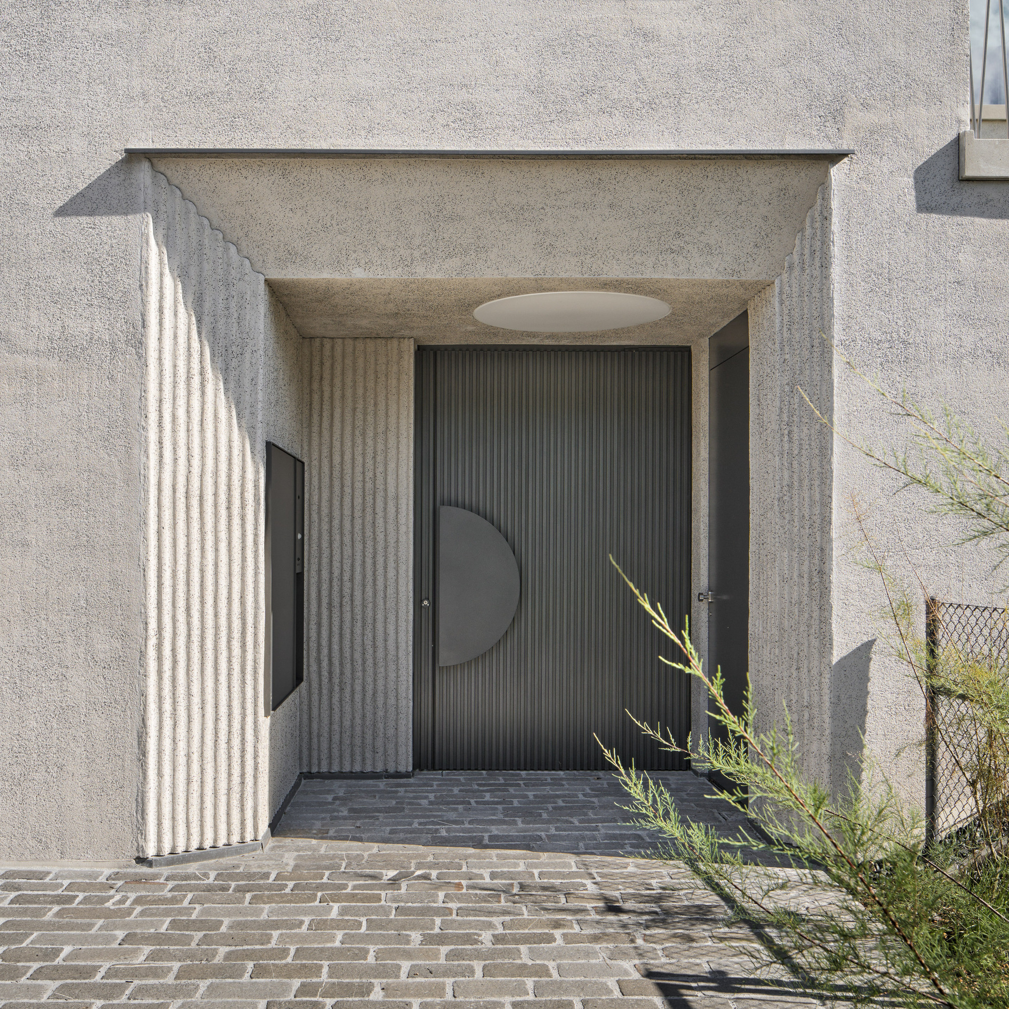 Recessed entry alcove with fluted concrete walls and dark ribbed door featuring a circular metal panel in sunlight