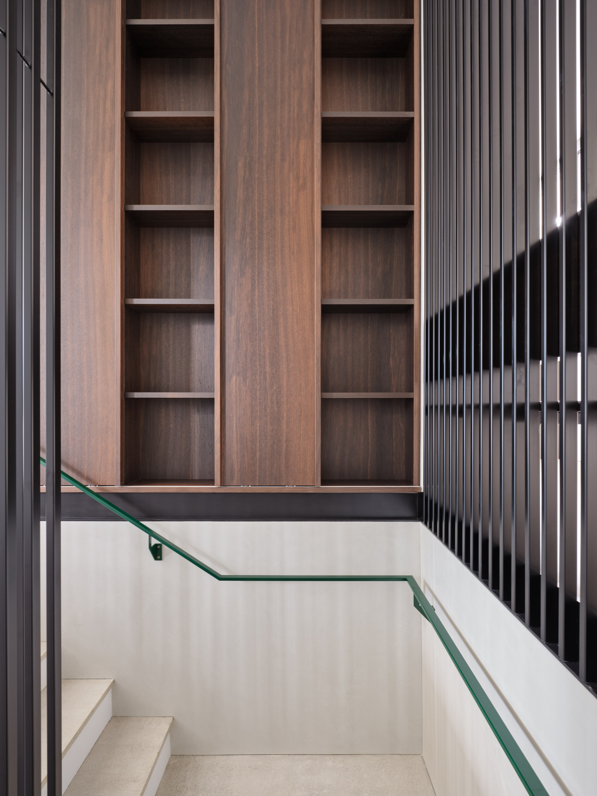 Stairwell with pale stone steps, walnut shelving and black vertical slat screen with green handrail