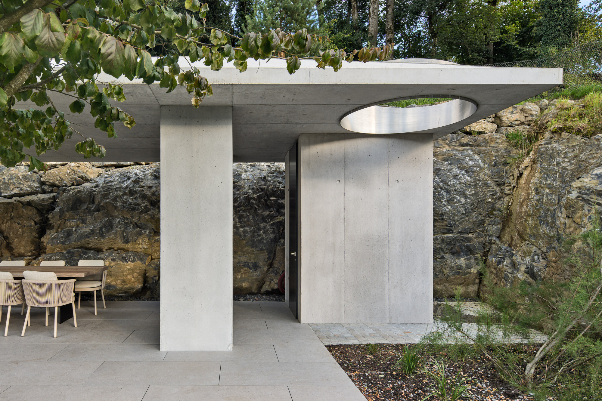 Concrete pavilion with flat roof and circular skylight opening set against exposed rock outcrop