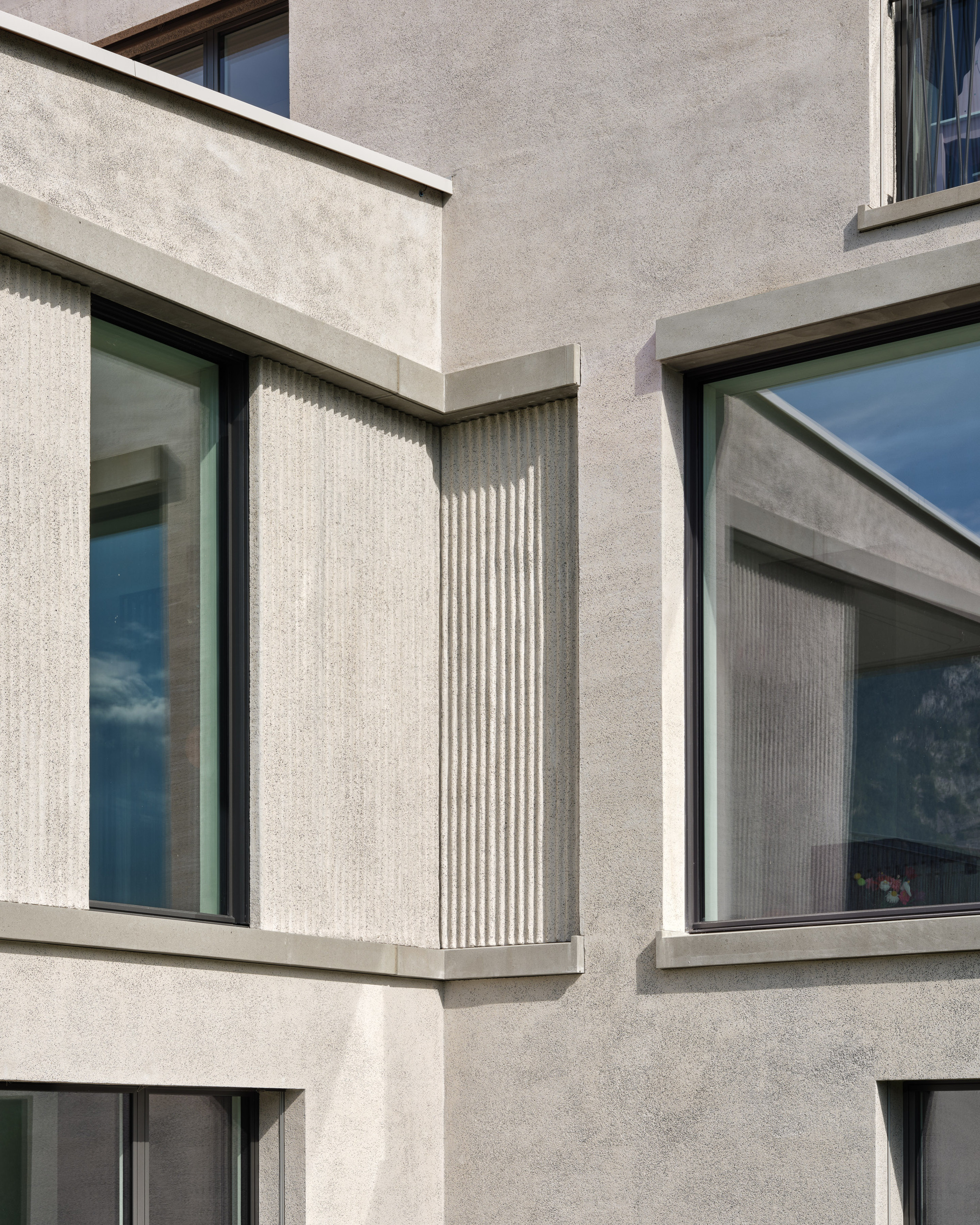 Exterior corner with fluted concrete panels between large windows reflecting sky and clouds