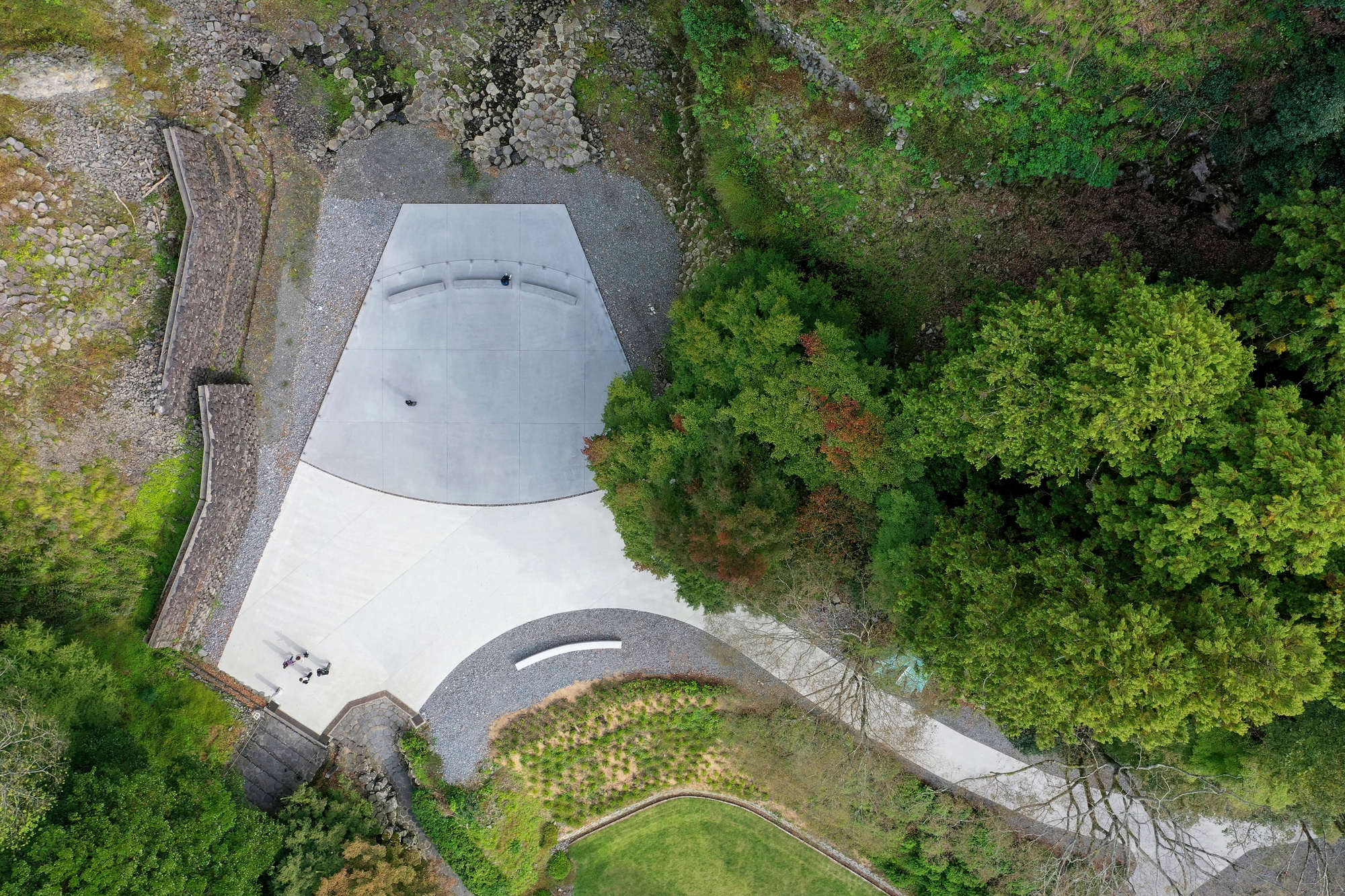 Aerial view of the curved concrete roof nestled among dense green forest and gravel paths