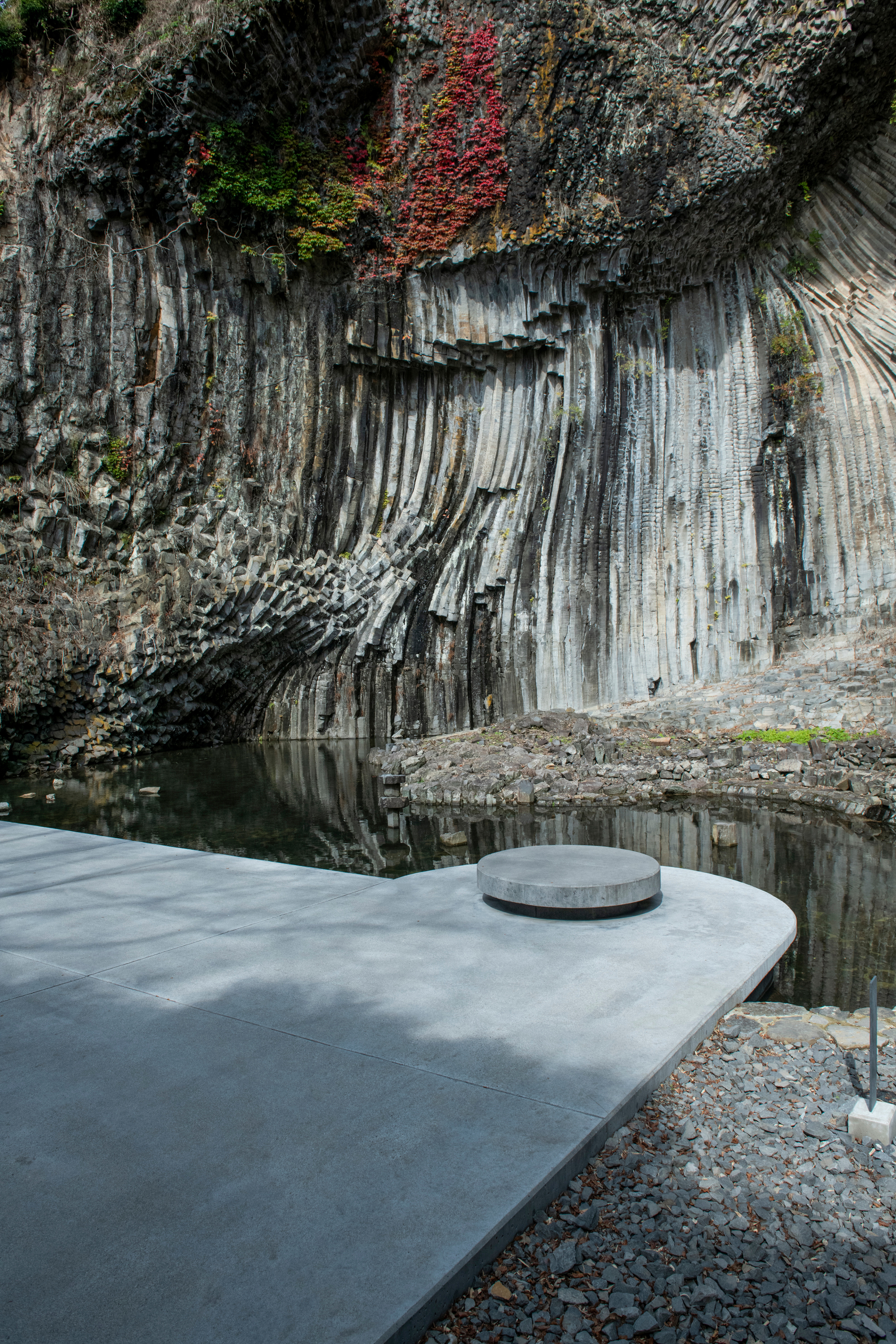 Curved concrete terrace with circular vent beside a still pool reflecting the columnar basalt cliff