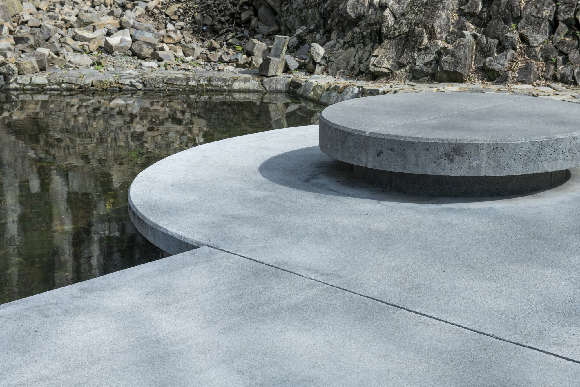 Layered circular concrete platforms beside a reflecting pool with quarried rock wall beyond