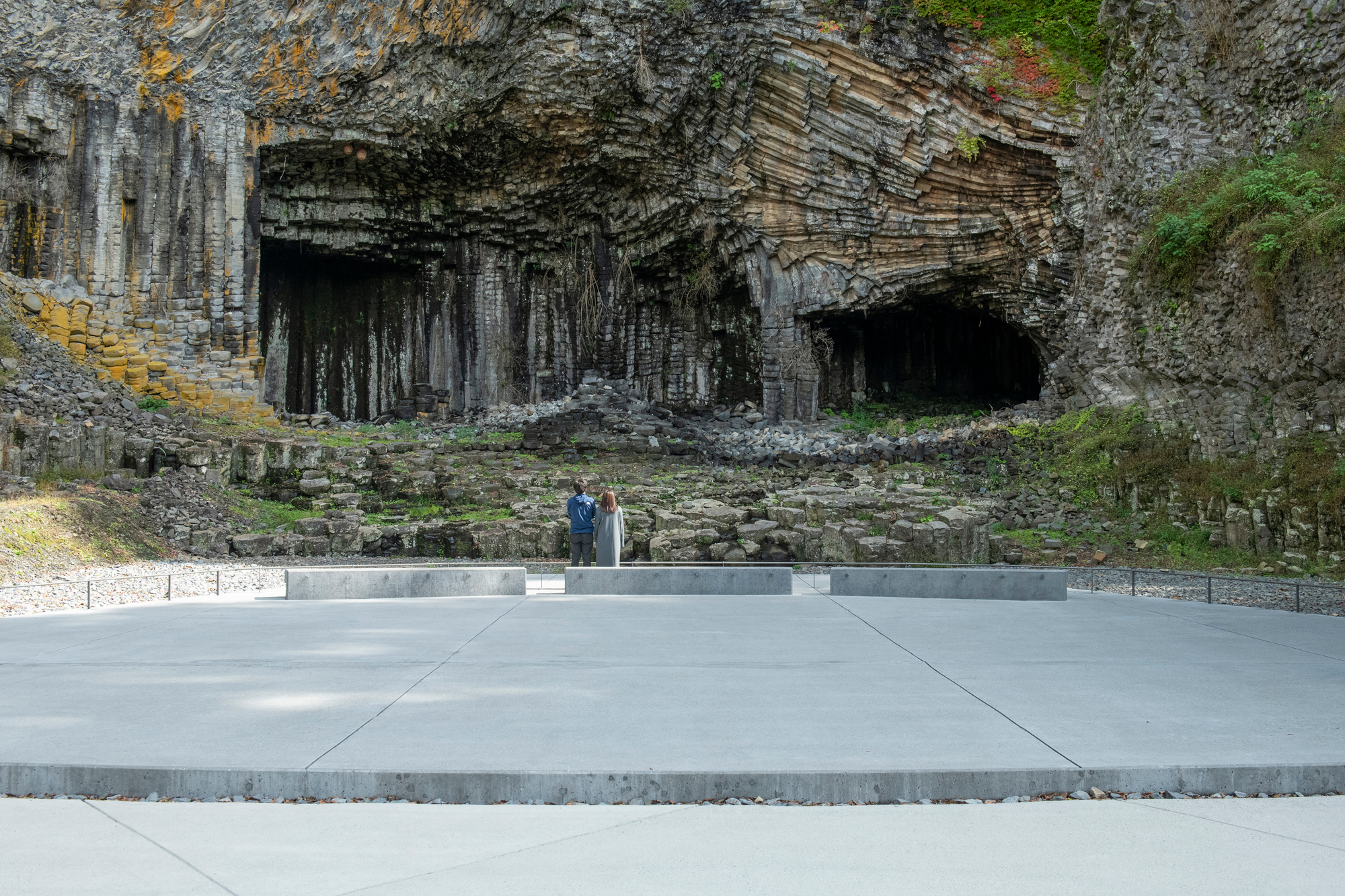 Concrete plaza with two visitors standing before the basalt cave openings and moss-covered walls