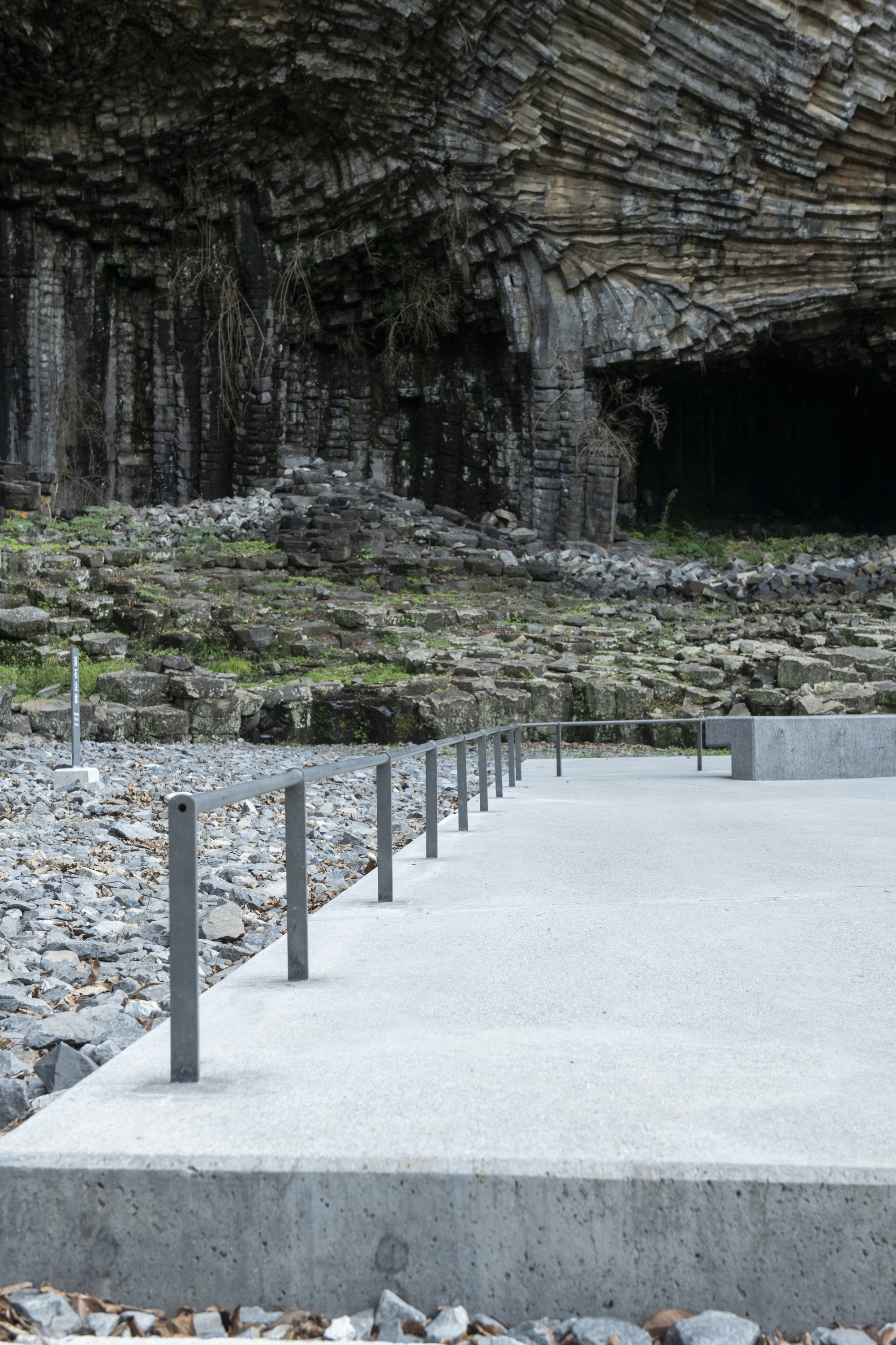 Concrete boardwalk with metal posts threading through a rocky shoreline below columnar basalt cliffs