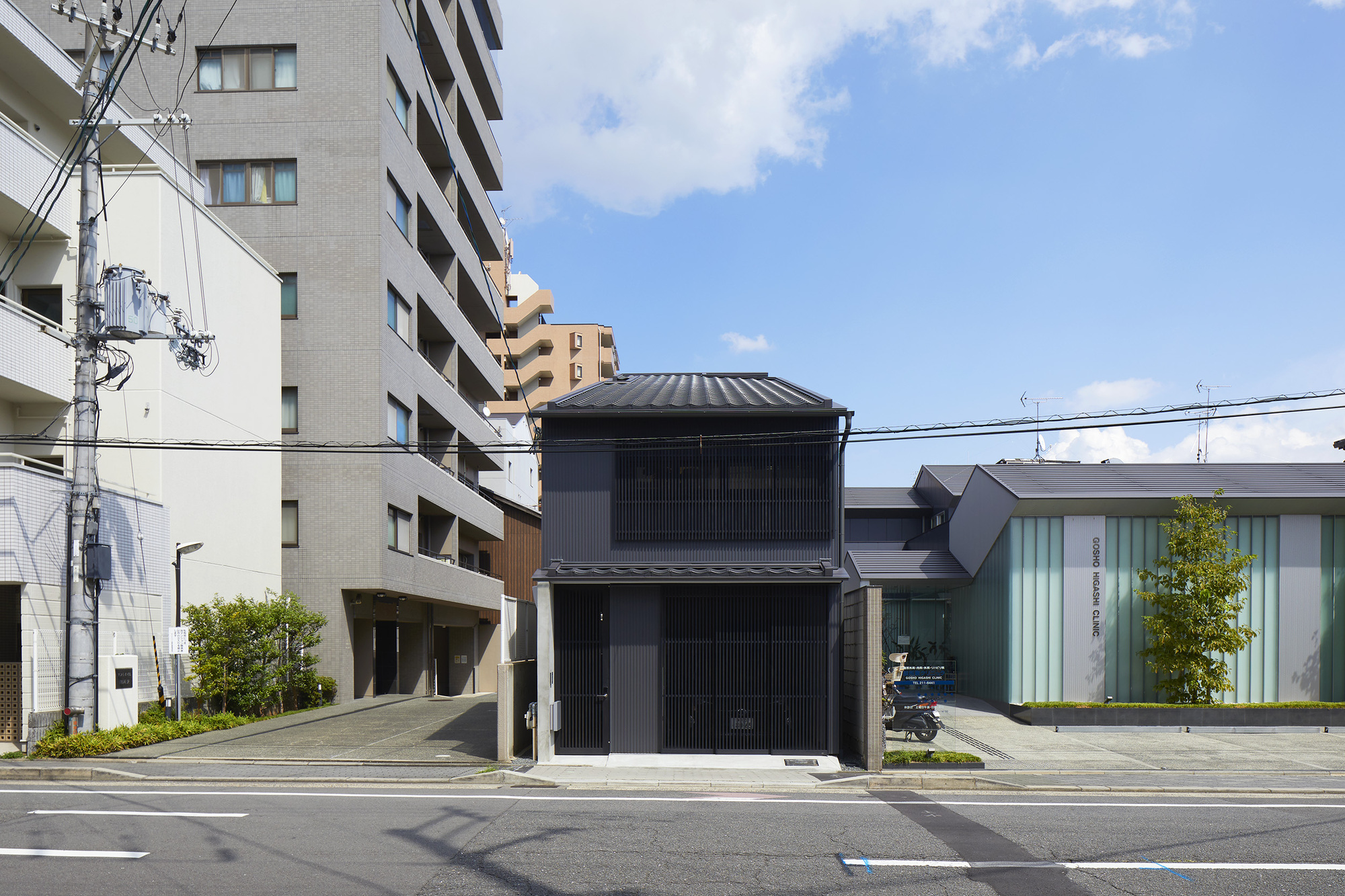 Street view of the vertical slatted black timber facade with tiled roof under scattered clouds