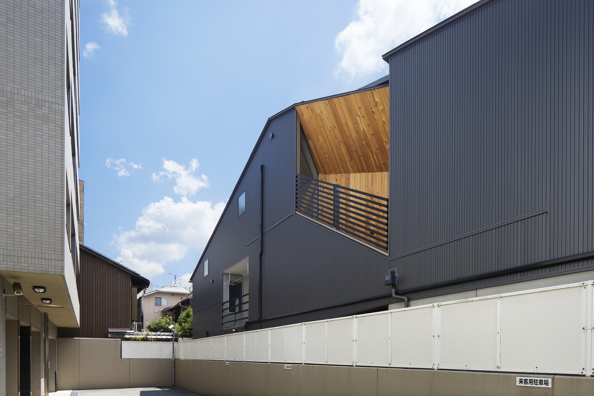 Dark corrugated metal facade with timber-lined balcony opening above a cream-colored perimeter wall under cloudy skies