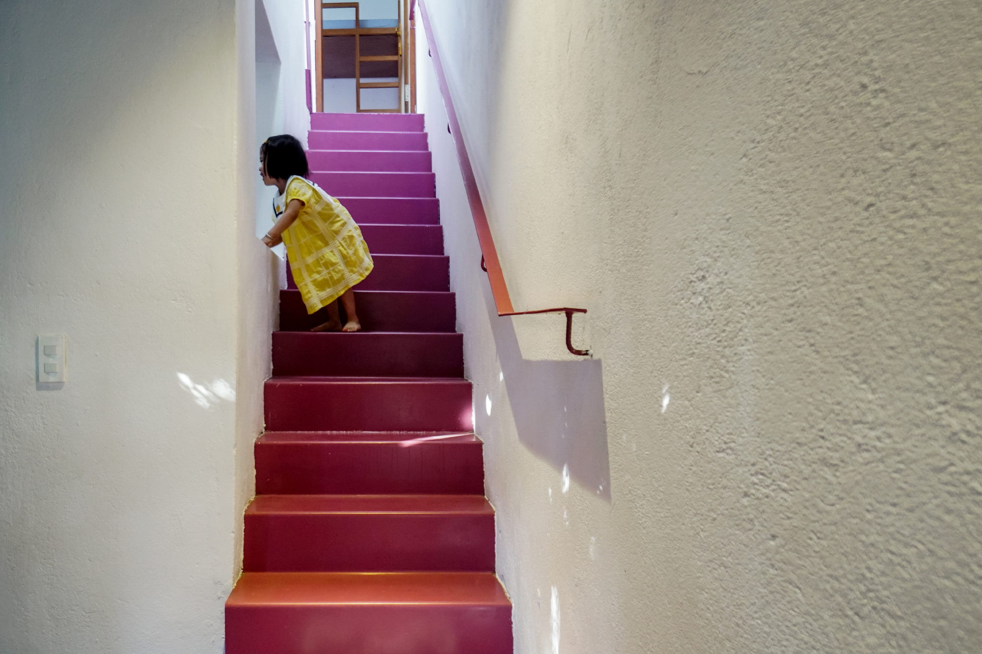 Child in a yellow dress ascending the maroon-painted staircase between white textured walls