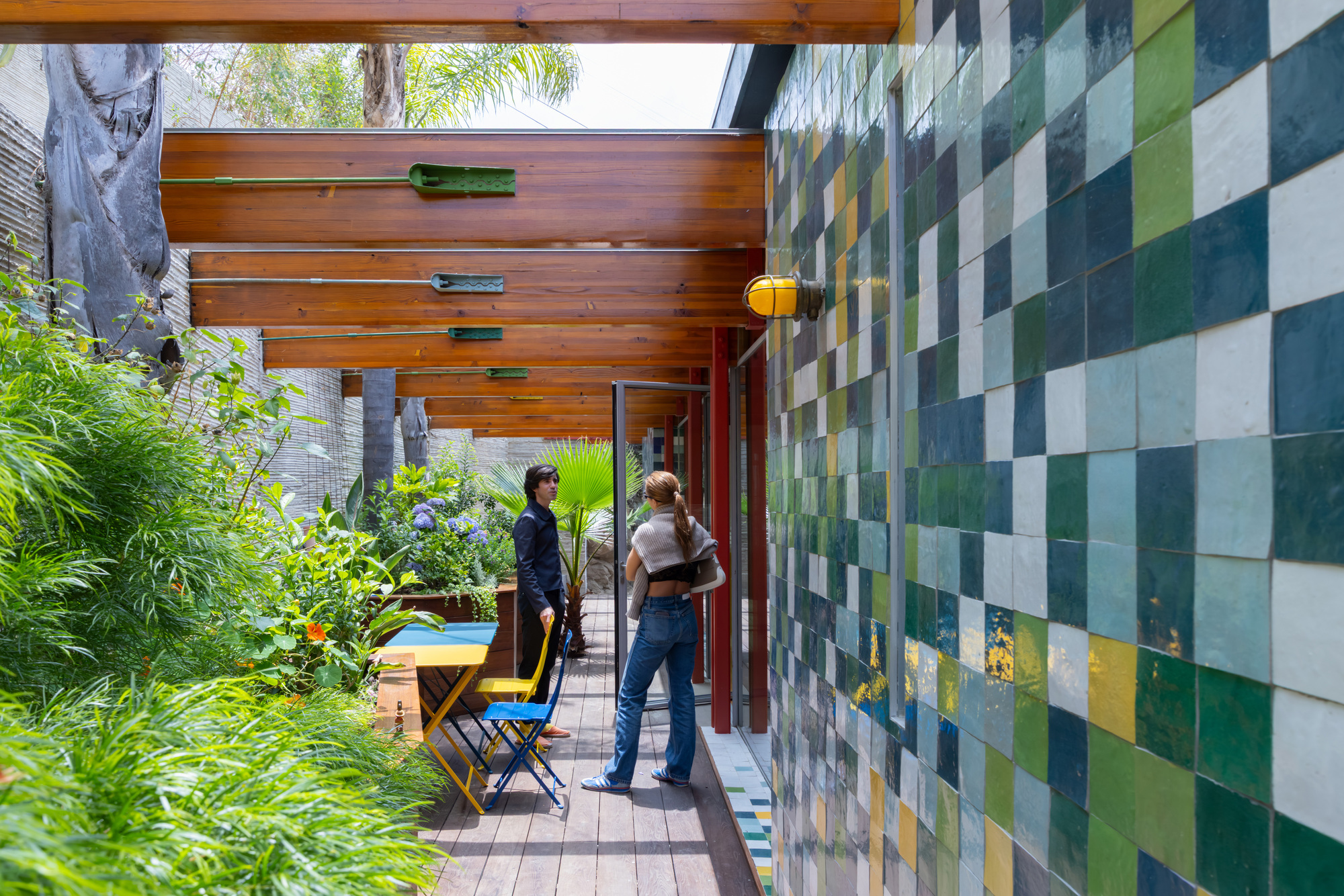 Covered walkway with multicolored glazed tile wall and timber ceiling overlooking a planted courtyard with two people