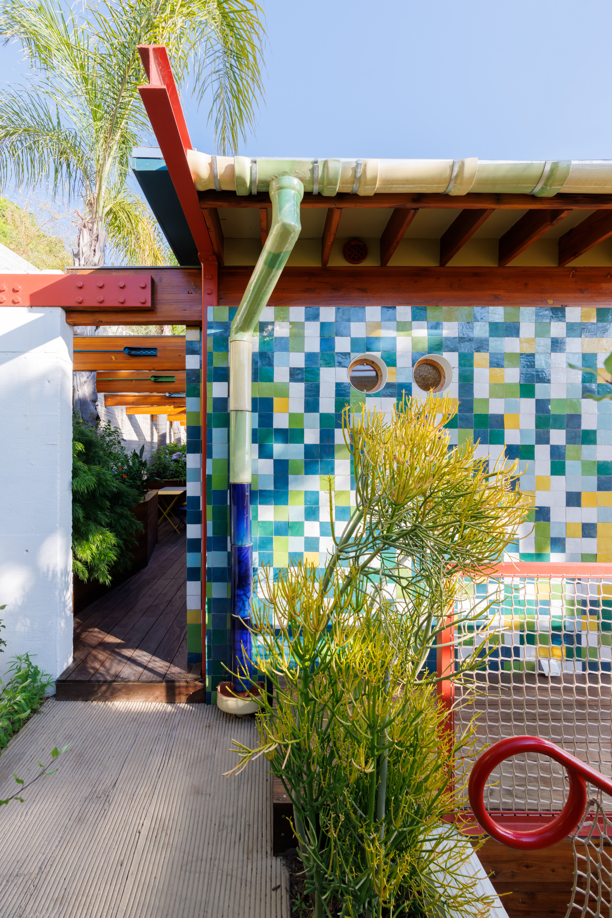Outdoor shower with multicolored mosaic tile wall and red steel pergola beside a palm tree
