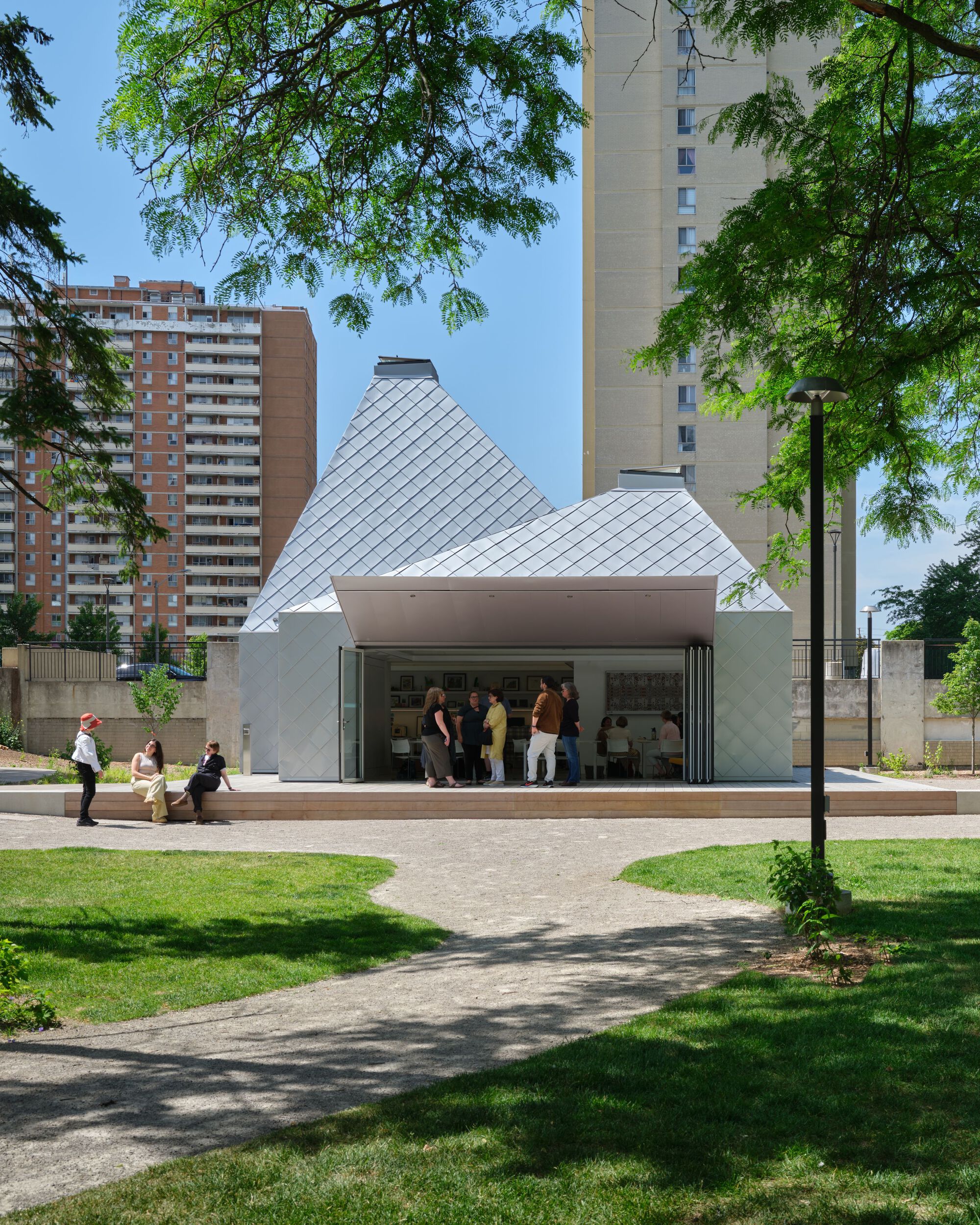 Long view across the lawn showing the pavilion's faceted roofline and glass openings under summer foliage