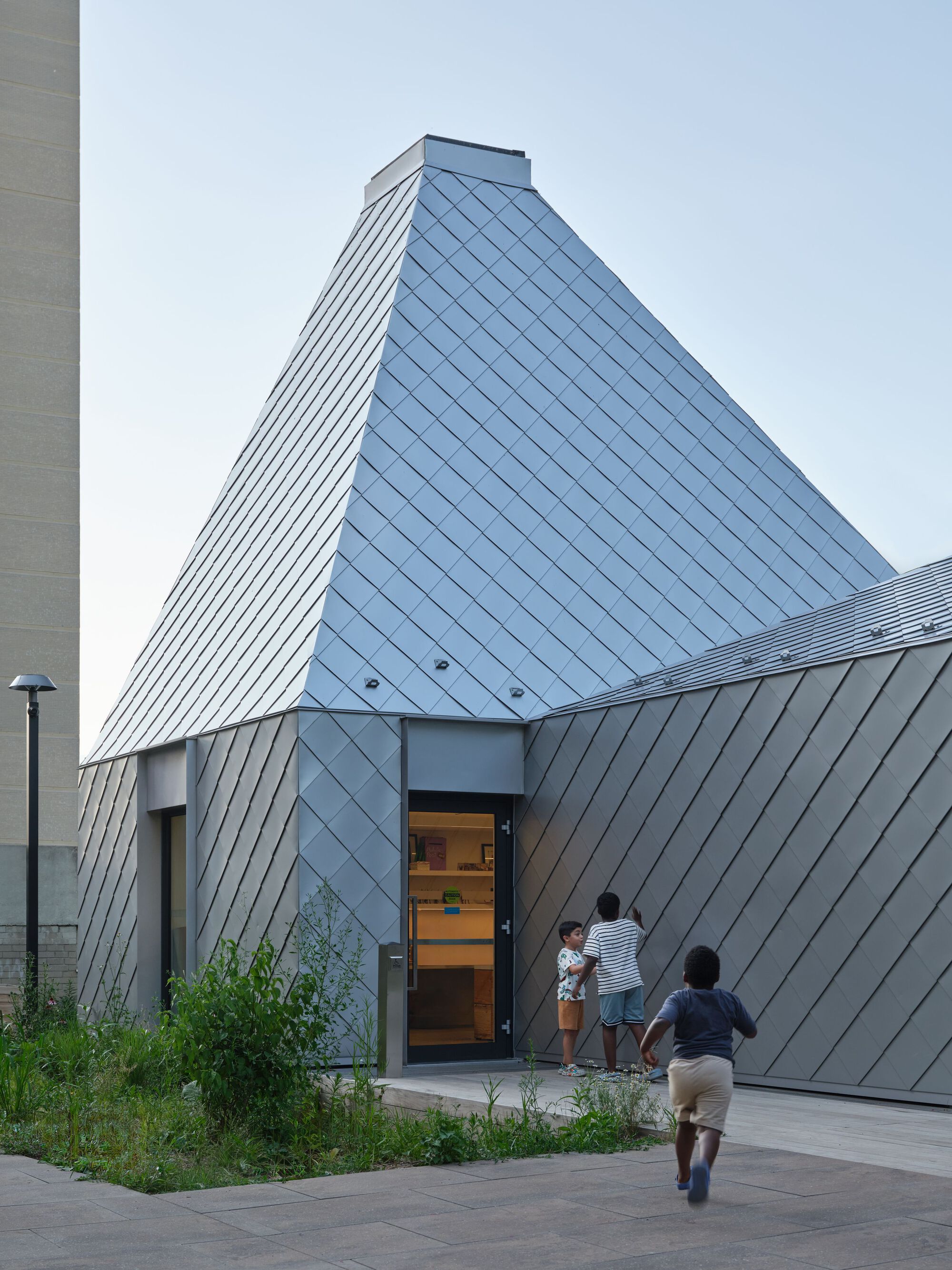 Diamond-patterned metal panel facade rising to a pyramidal volume as children run toward the entrance