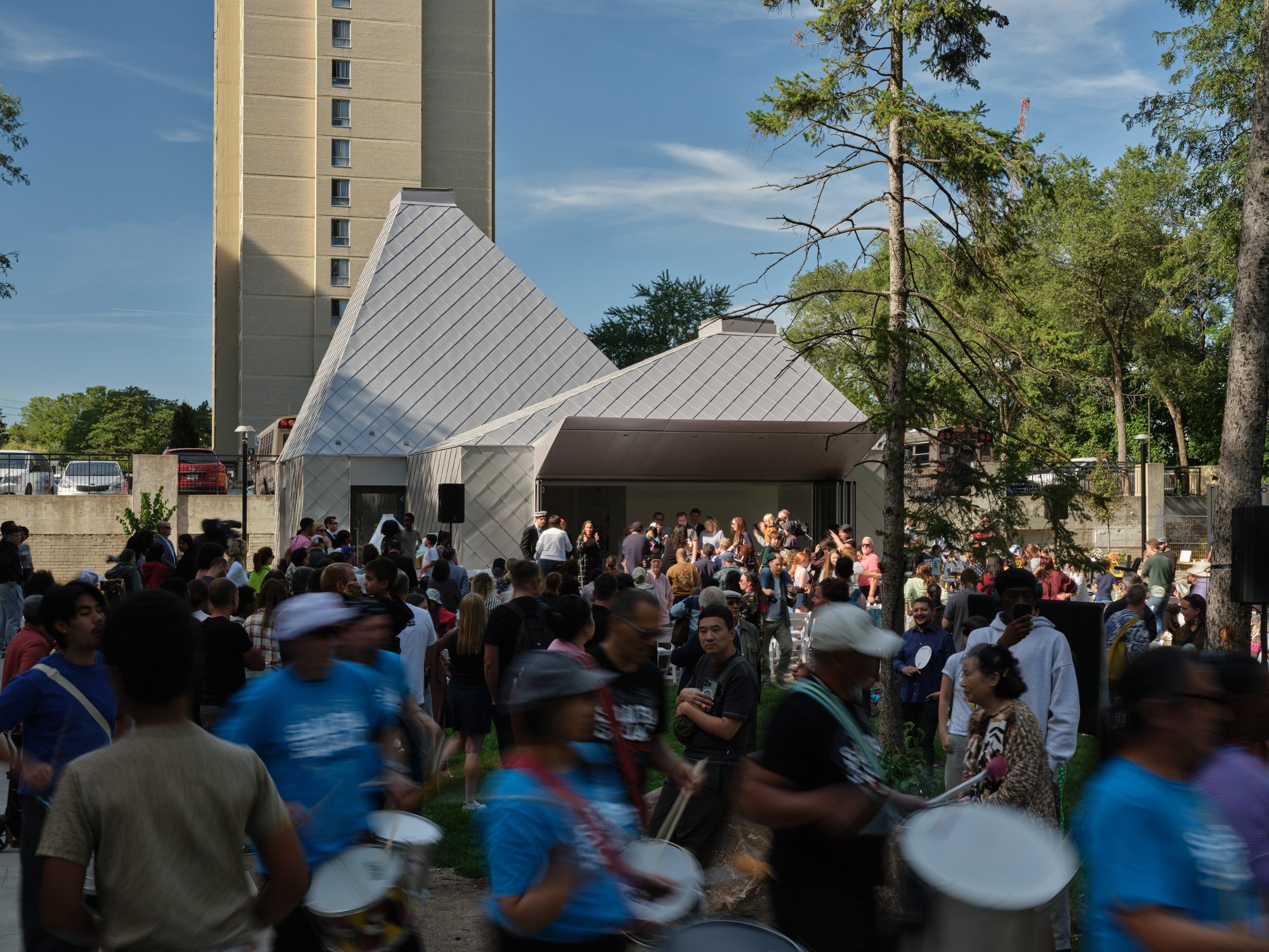 Gathered crowd surrounds the gray metal-clad pyramidal volumes in the afternoon light