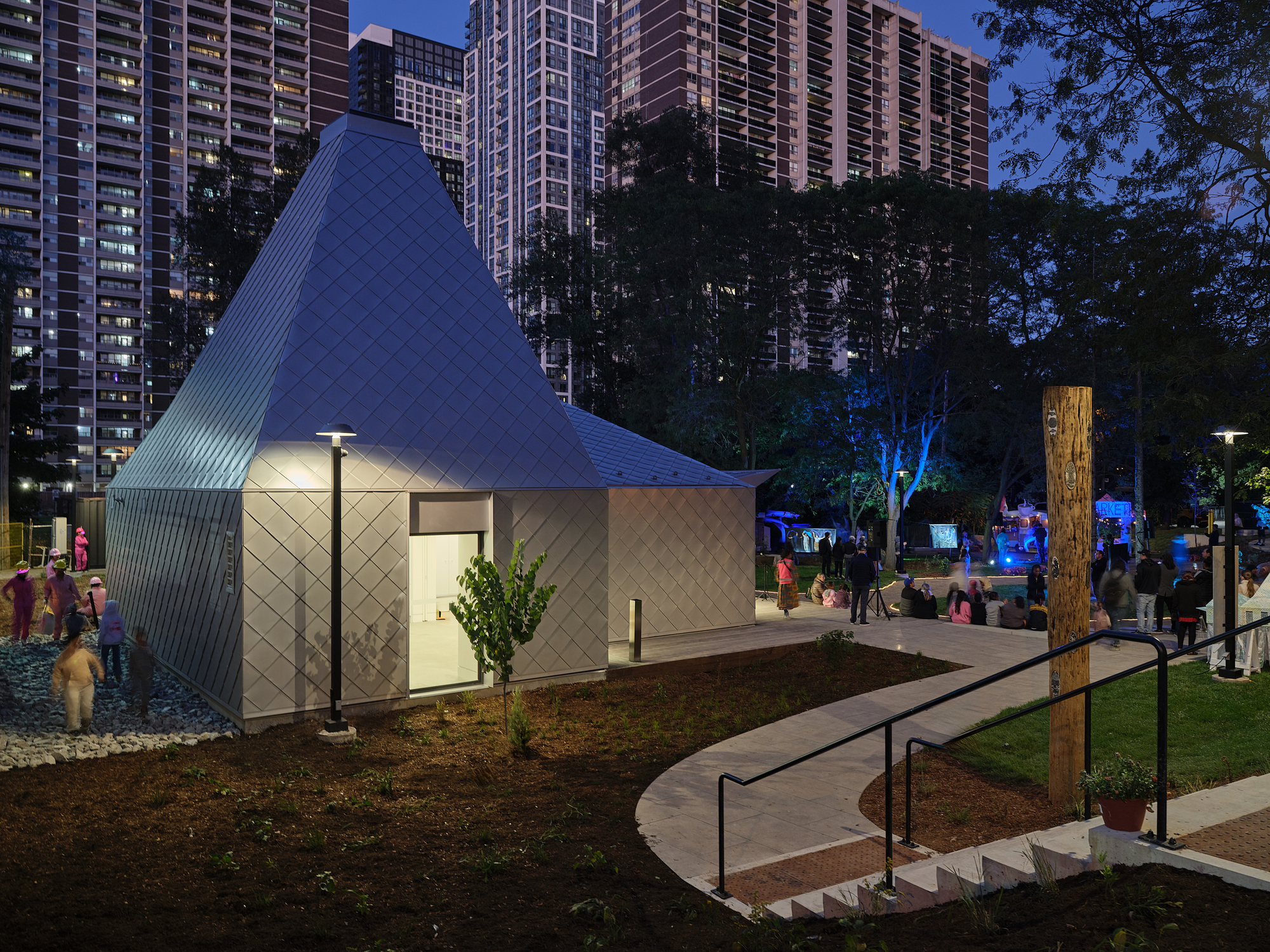 Illuminated pavilion with diamond-pattern metal facade and glazed openings at twilight among residential towers