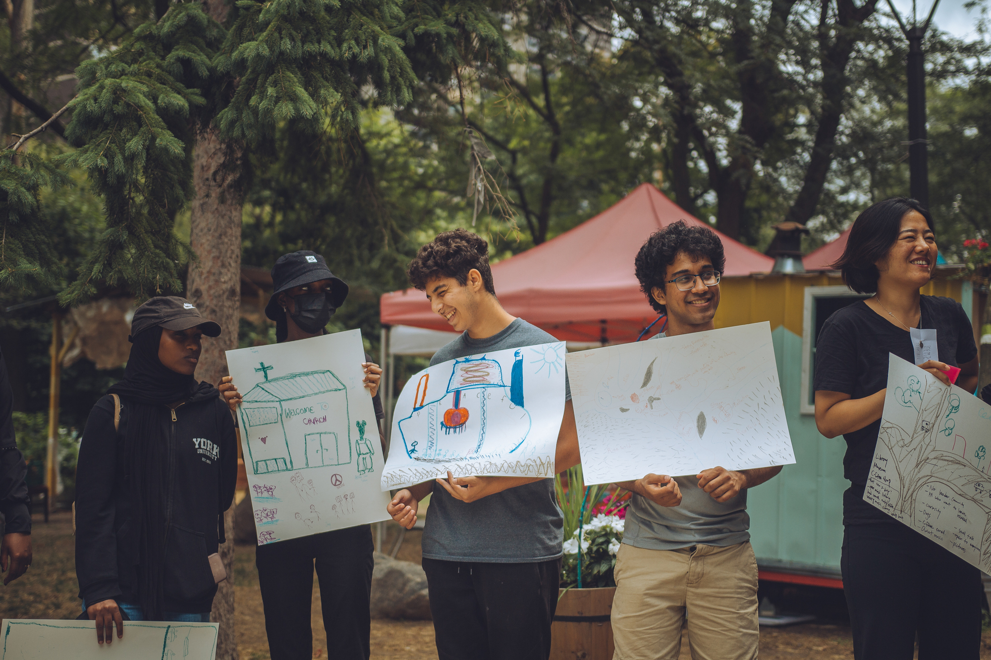 Group of participants holding architectural drawings outdoors beneath red canopy tents and mature trees