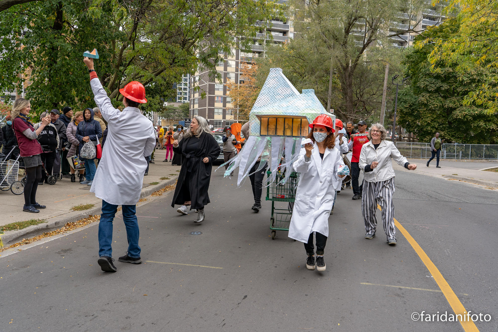 Street parade with costumed participants wheeling paper model with checkered pyramidal roof through autumn leaves