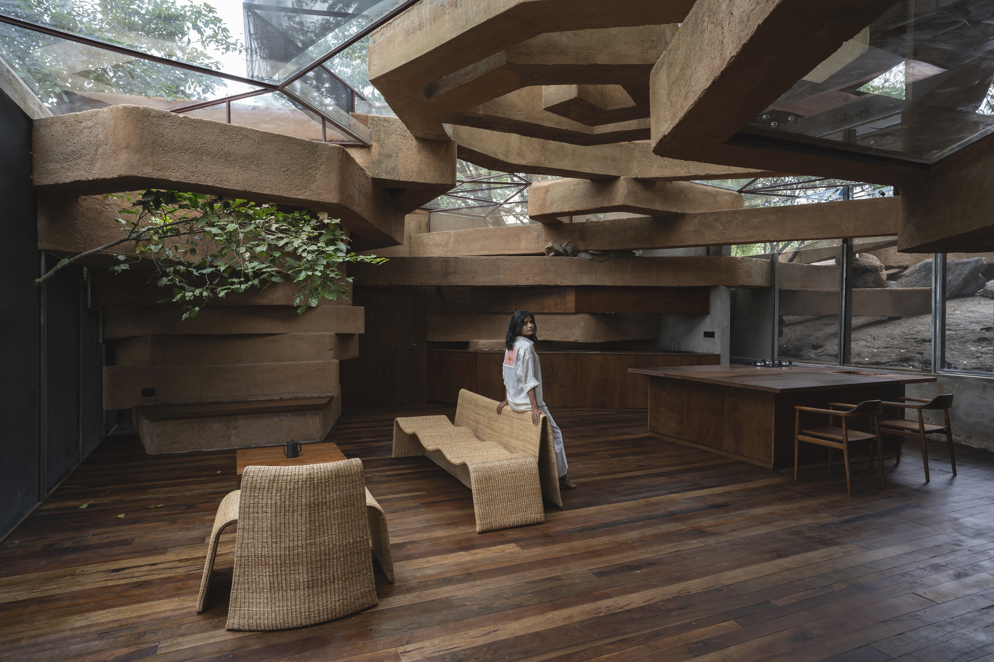 Interior living space with stacked rammed earth beams overhead and a woman standing among woven seating