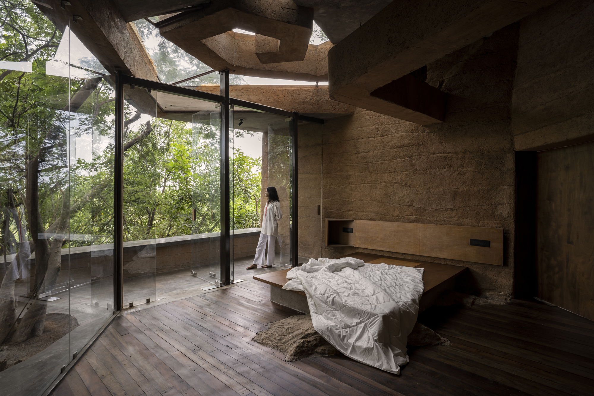 Bedroom with floor-to-ceiling glass walls opening to trees and the adjacent rammed earth wall