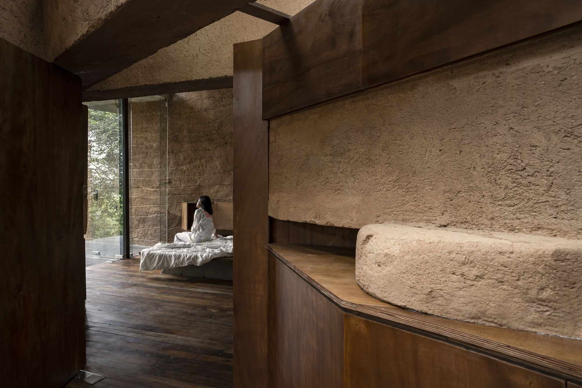Curved wooden millwork meeting textured earthen walls with a figure seated on a bed beyond