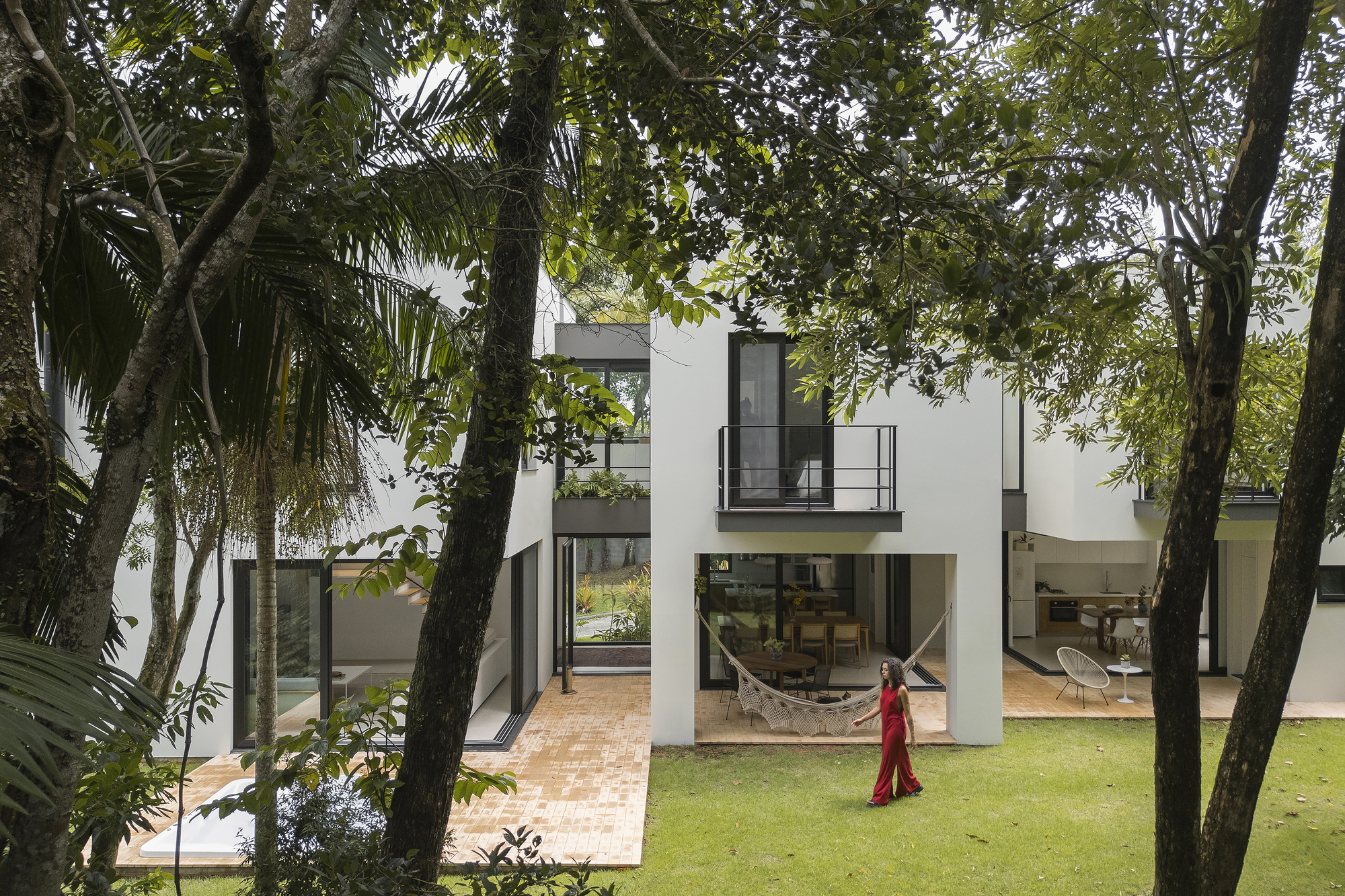 Garden courtyard view with person in red dress walking across lawn between white volumes and mature trees