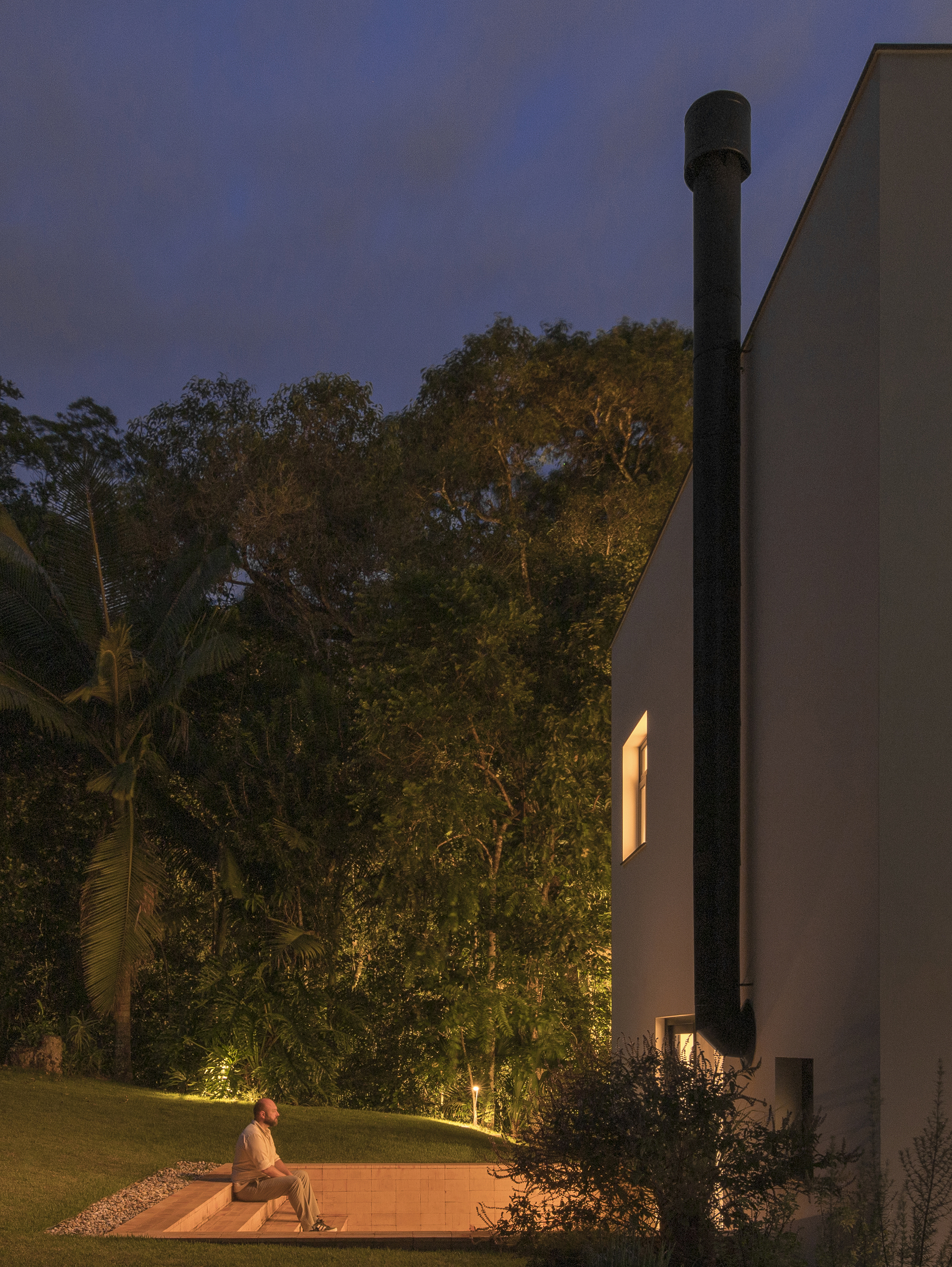 Black cylindrical flue rising against the white facade with a figure seated on timber deck at dusk