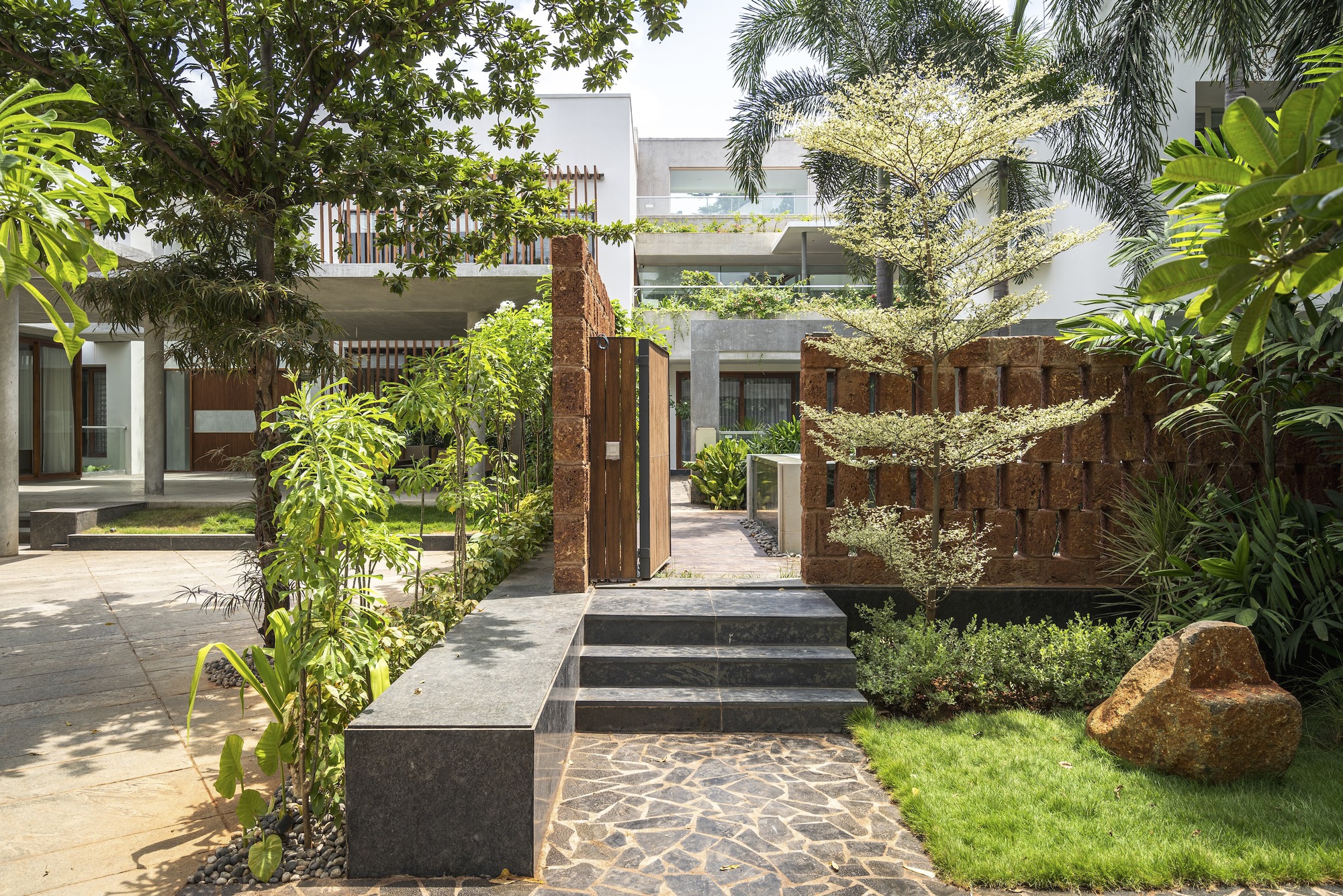 Garden courtyard with textured red stone wall, concrete steps, and planted beds beneath palm trees