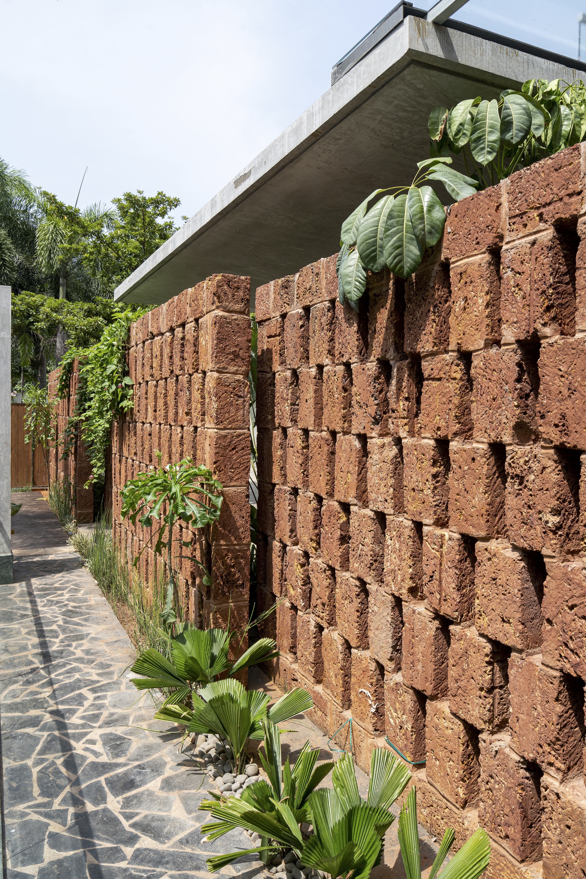 Perforated red volcanic block wall with concrete canopy above and tropical planting along the paved walkway