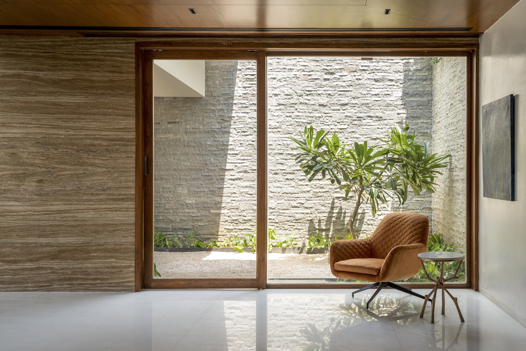 Interior view through timber-framed glazing to a planted courtyard with textured stone walls in daylight
