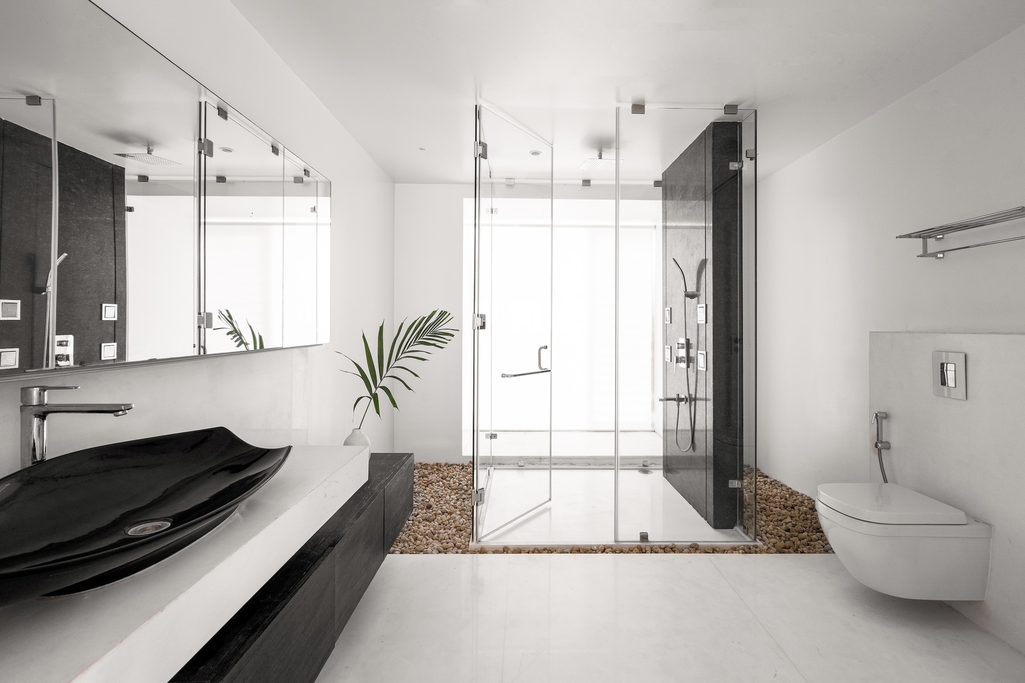Bathroom interior with black vessel sink, glass-enclosed shower on river stones, and palm frond in background
