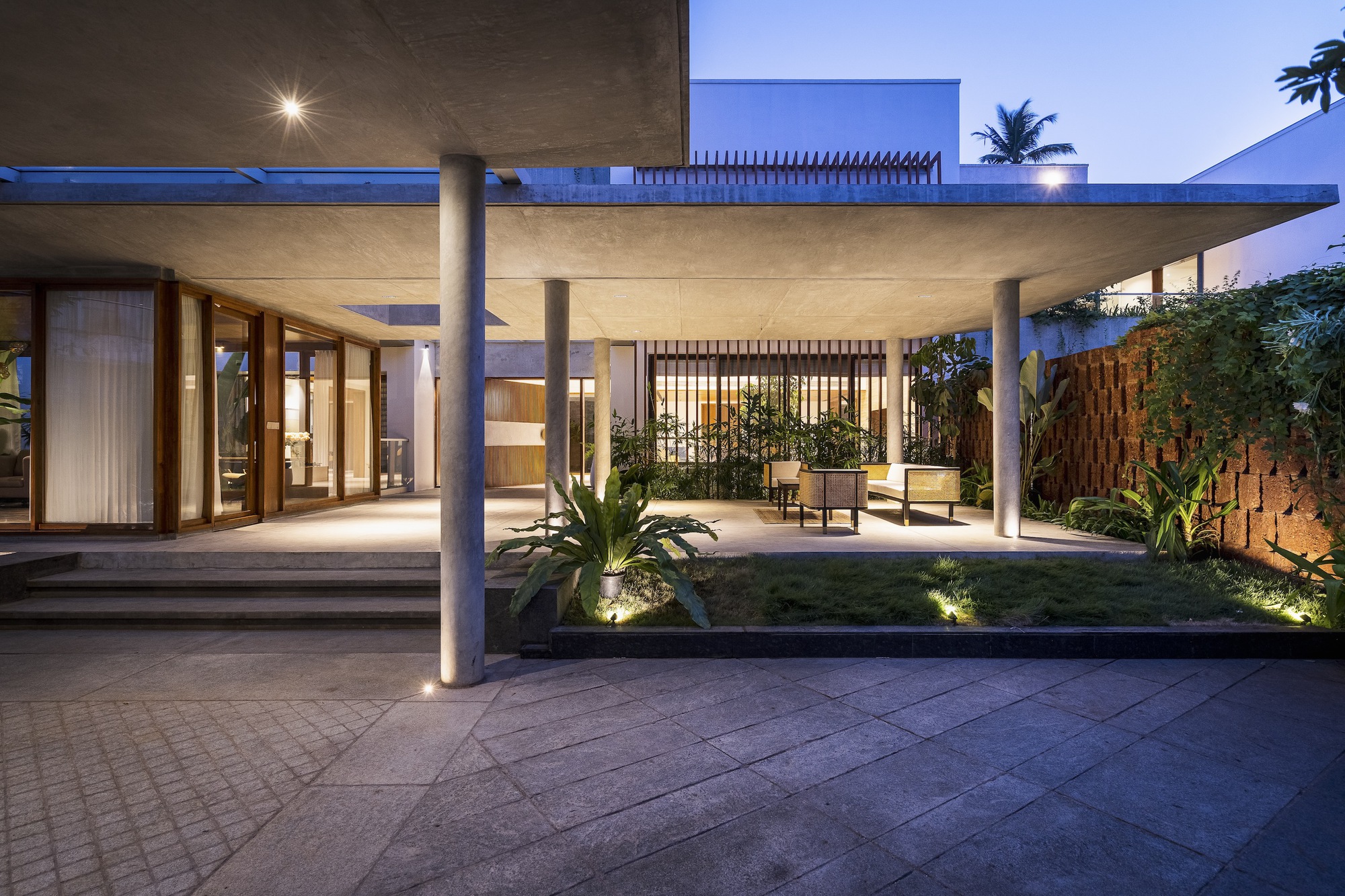 Covered outdoor terrace with concrete columns and slab roof at dusk, illuminated by uplights and tropical plantings