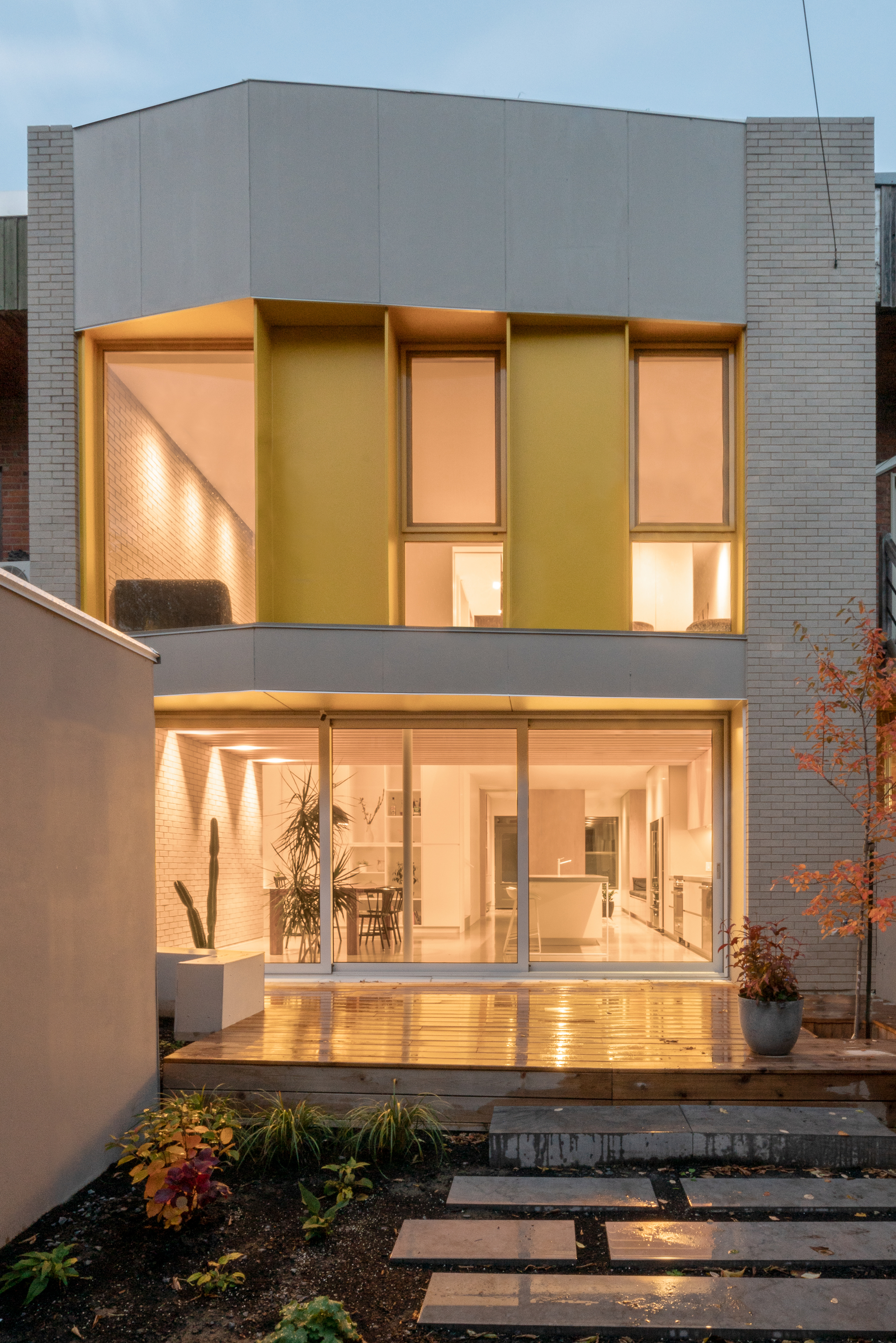 Rear garden facade with warm-lit glazing and timber deck stepping down to planted beds at dusk