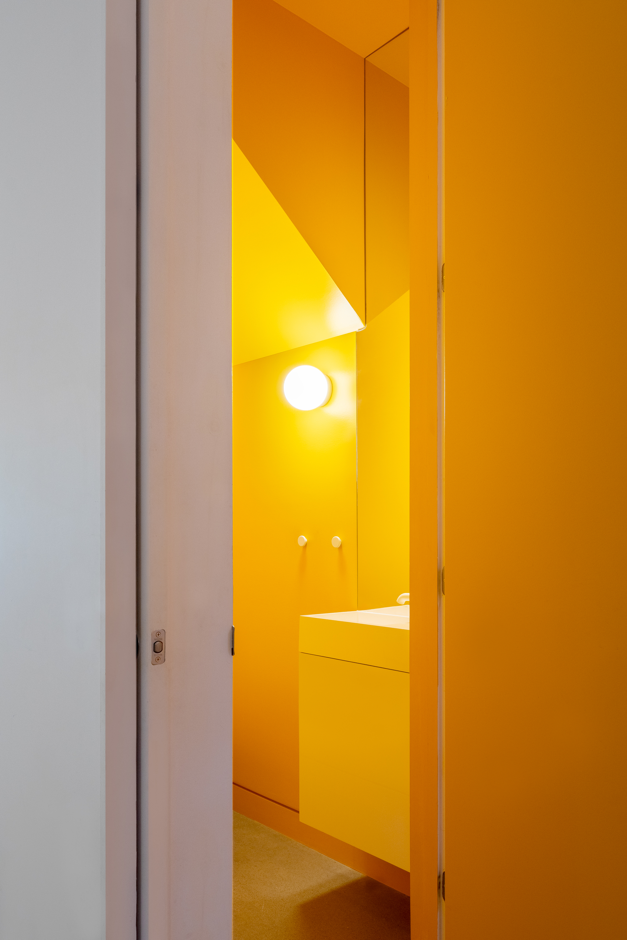 View through a narrow doorway into a fully tiled bathroom with yellow surfaces and circular wall sconce