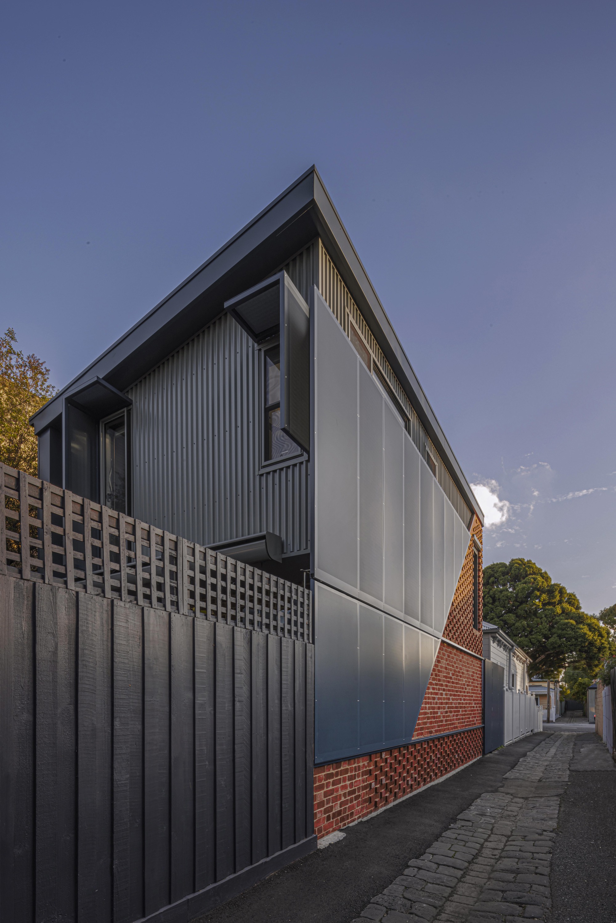 Laneway elevation showing the corrugated metal upper level and patterned brick base at dusk