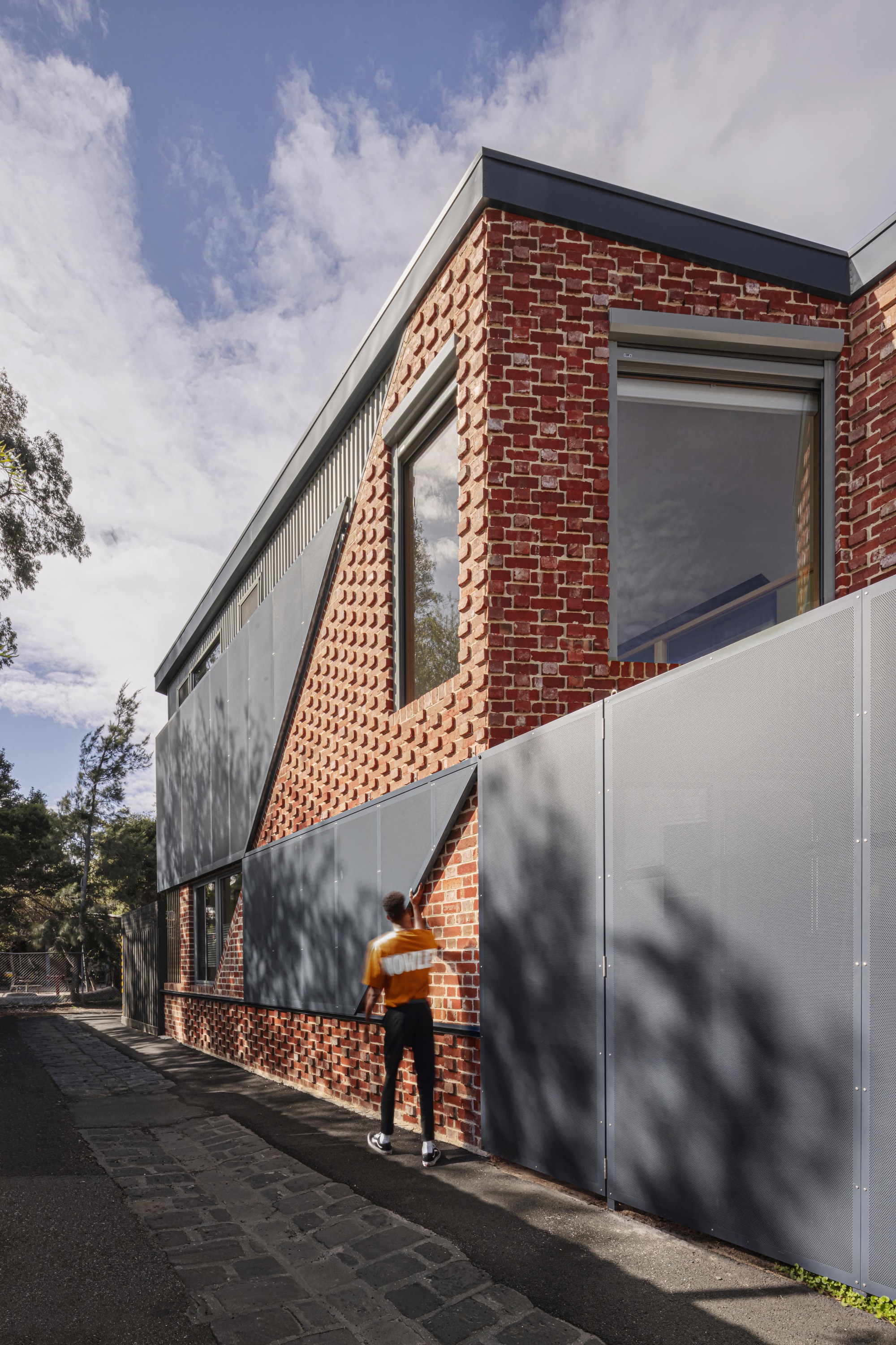 Pedestrian passing the perforated brick screen and metal gate along the tree-lined street