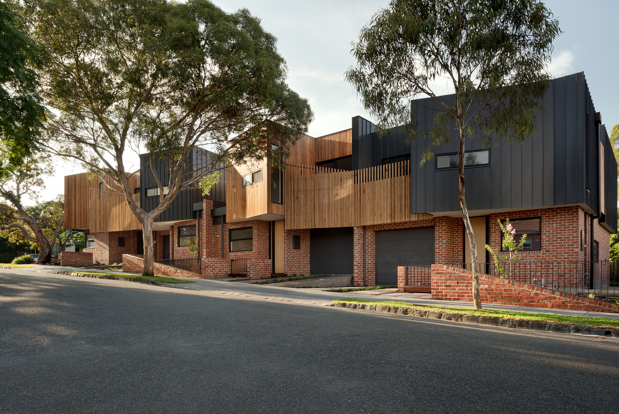 Street view of the multi-unit facade with red brick base, vertical timber cladding, and dark metal volumes beneath mature eucalyptus trees