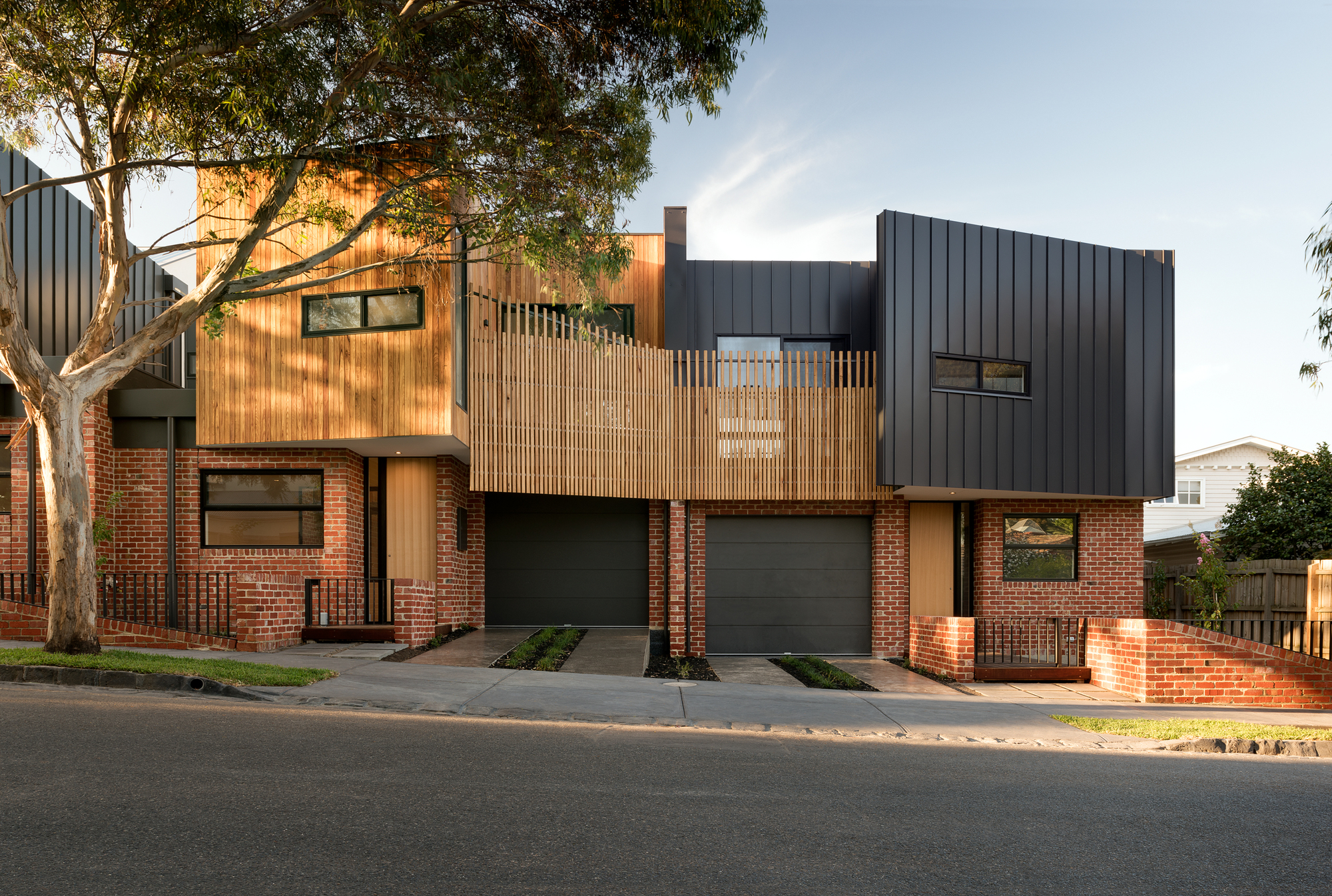 Row of garages with red brick piers and timber screening at dusk, with dark metal upper levels and gum trees overhead