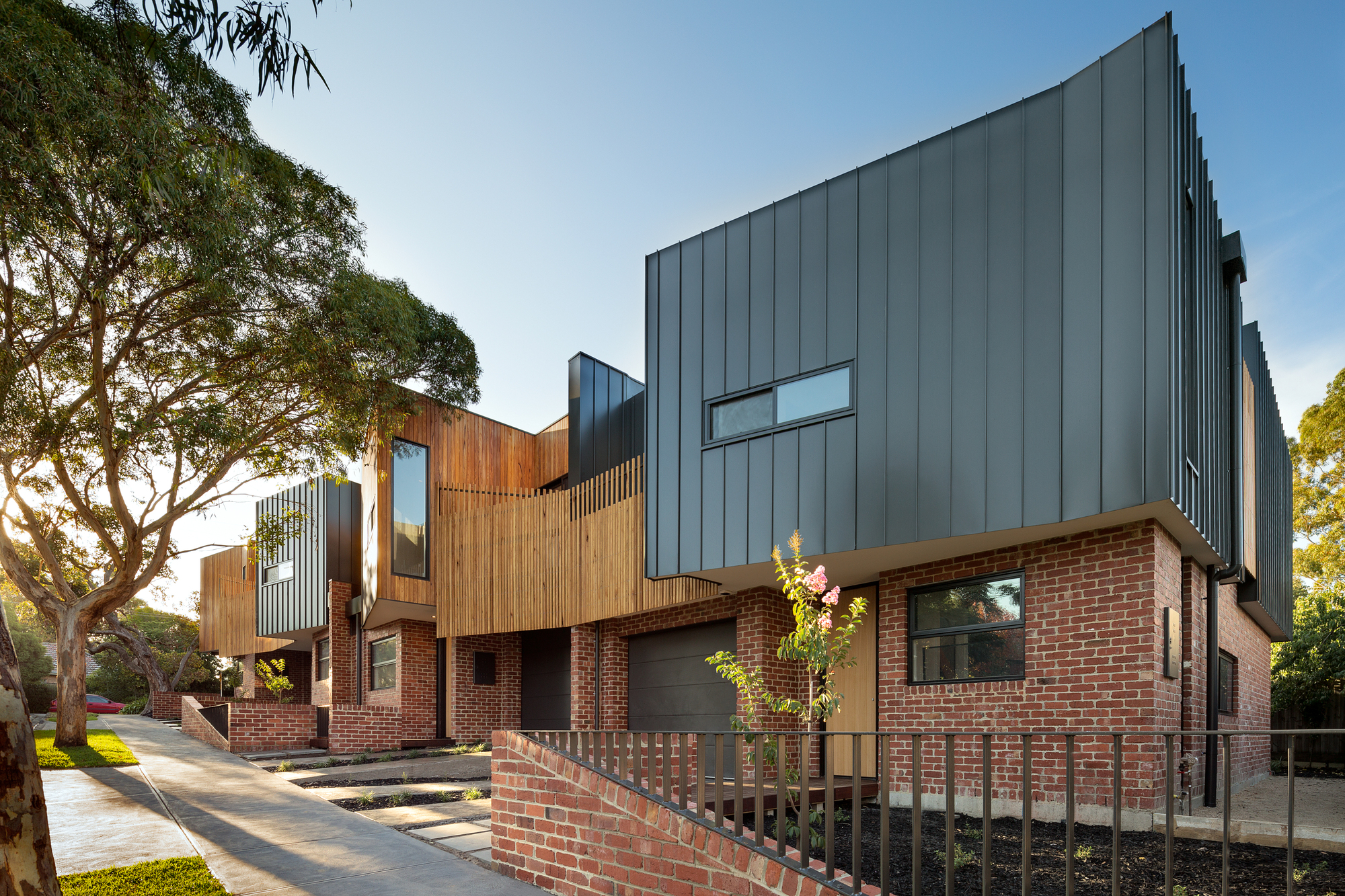 Street view of row housing with red brick base, timber cladding and charcoal metal volumes in afternoon light